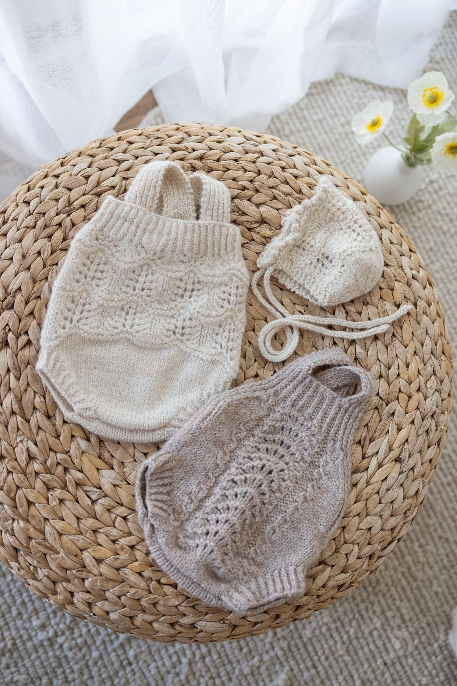 Cozy knitted baby rompers and booties in neutral beige and gray tones displayed on woven rattan mat with small flowers.