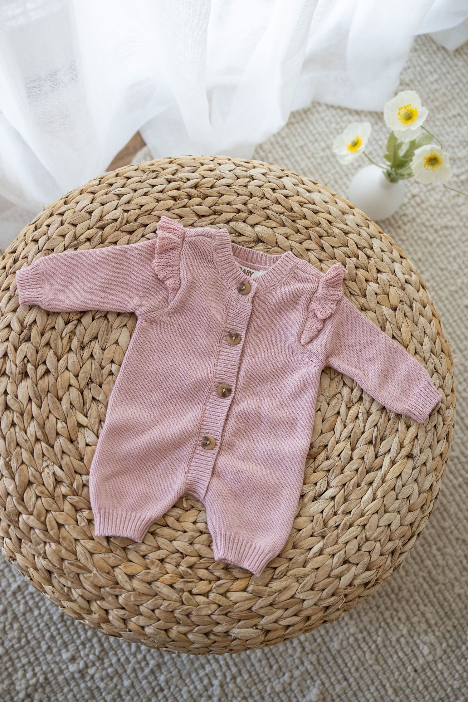 Pink knit baby romper with button front displayed on circular woven rattan mat with white daisies in background.