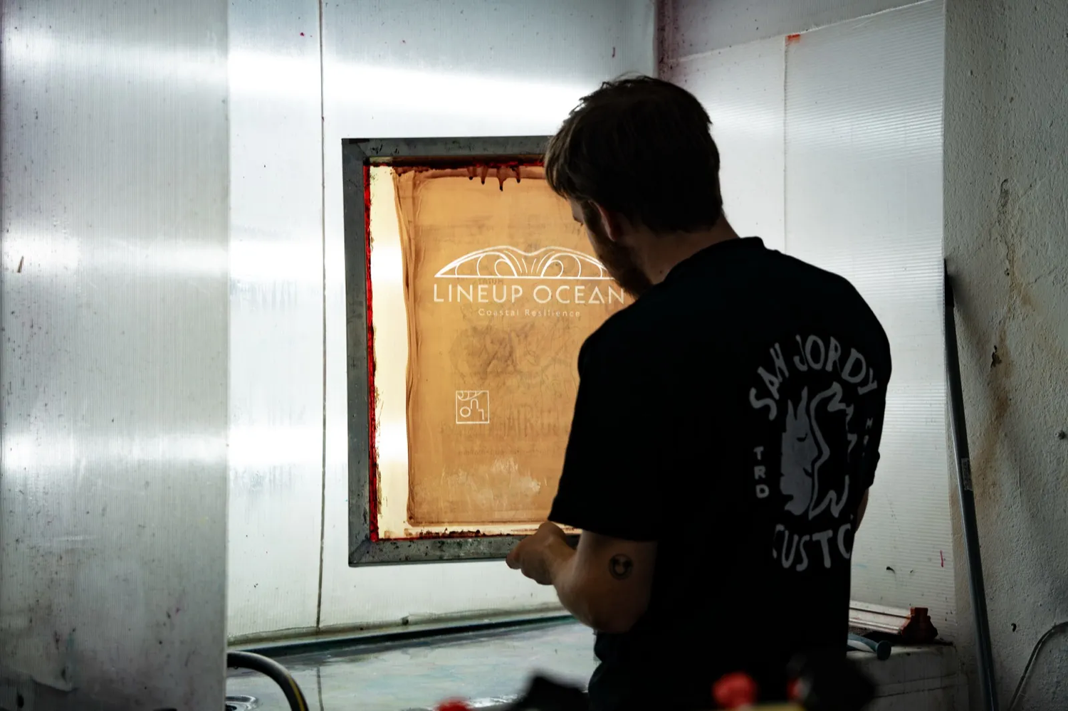 Impression artisanale sur cadre de sérigraphie dans un atelier, dos d’un artiste concentré.