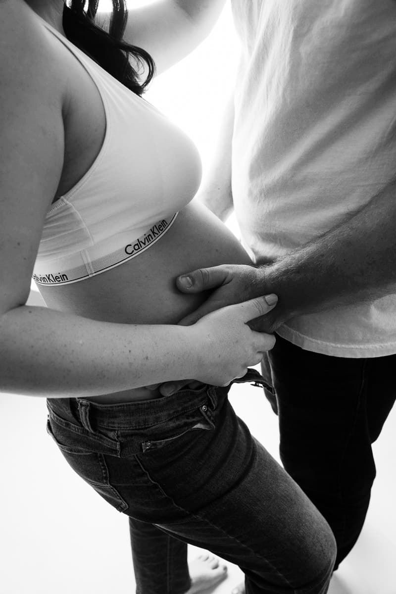 Black and white maternity portrait of a pregnant woman standing barefoot against a plain background, smiling gently while holding her belly