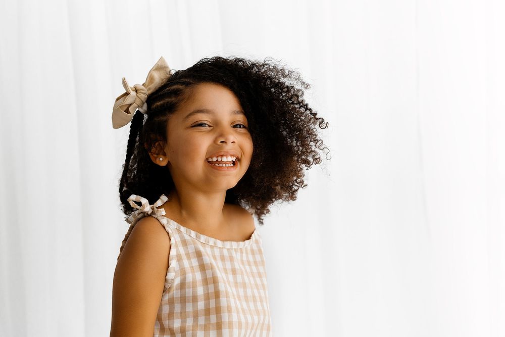 A young child in a gingham dress with curly natural hair and hair bows laughs joyfully against a white background.