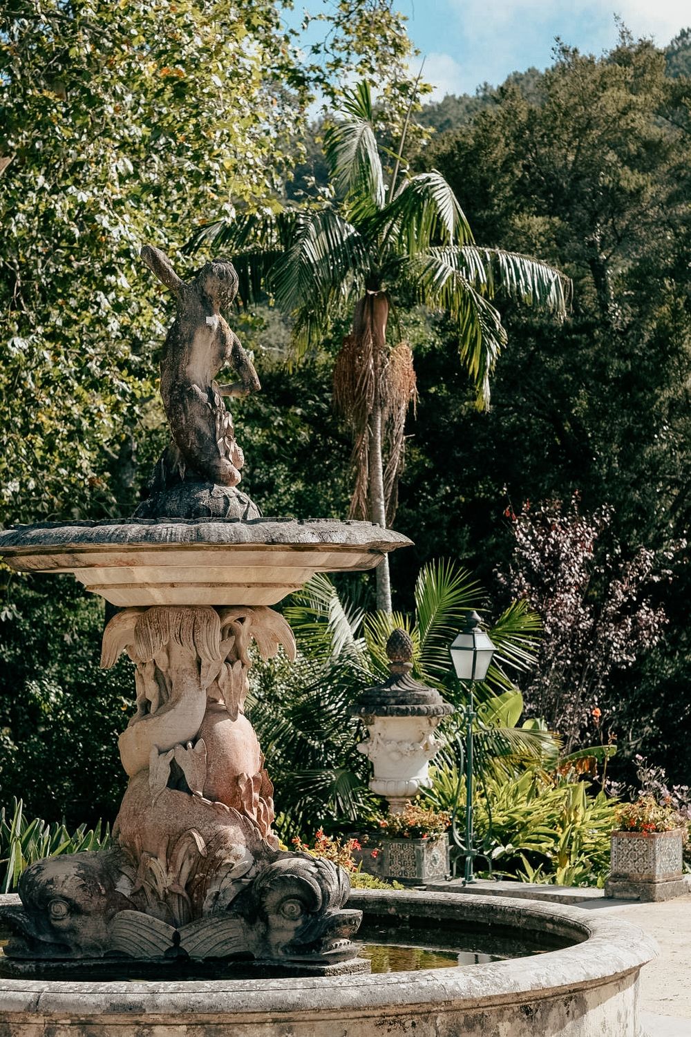 Stone garden statues and ornate architectural elements adorn a lush tropical landscape with palm trees and greenery.