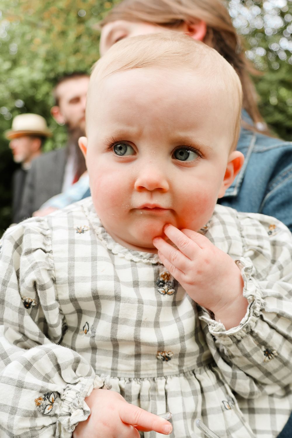 Baby wearing grey and white checkered dress with bright blue eyes looks pensively while touching their face.