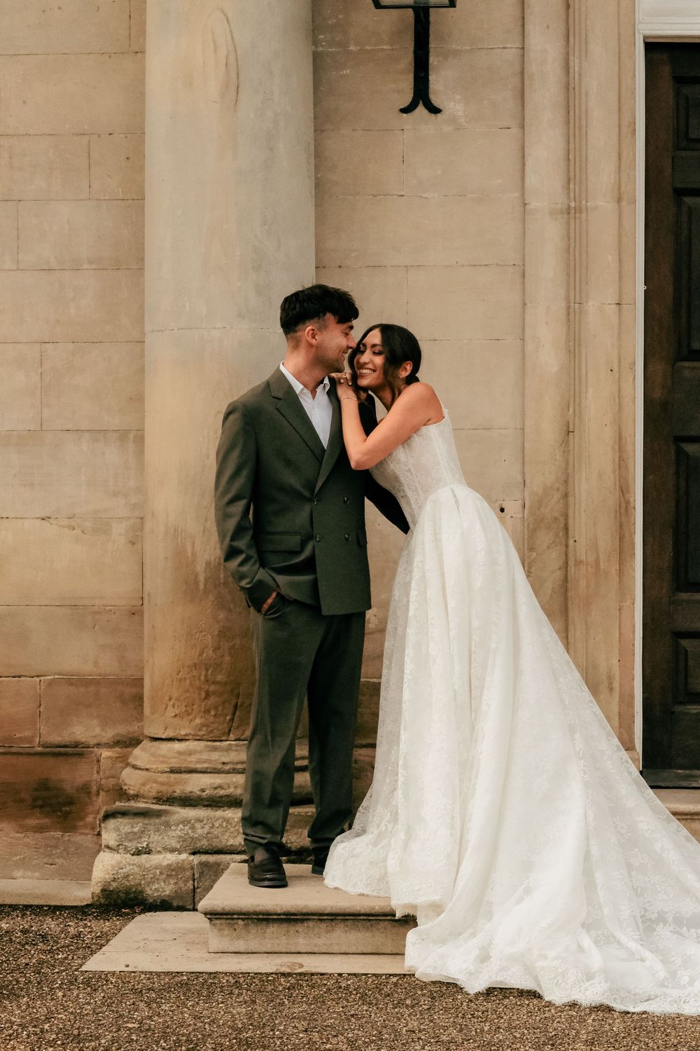 A romantic wedding couple shares intimate kisses in front of elegant stone pillars at their venue.