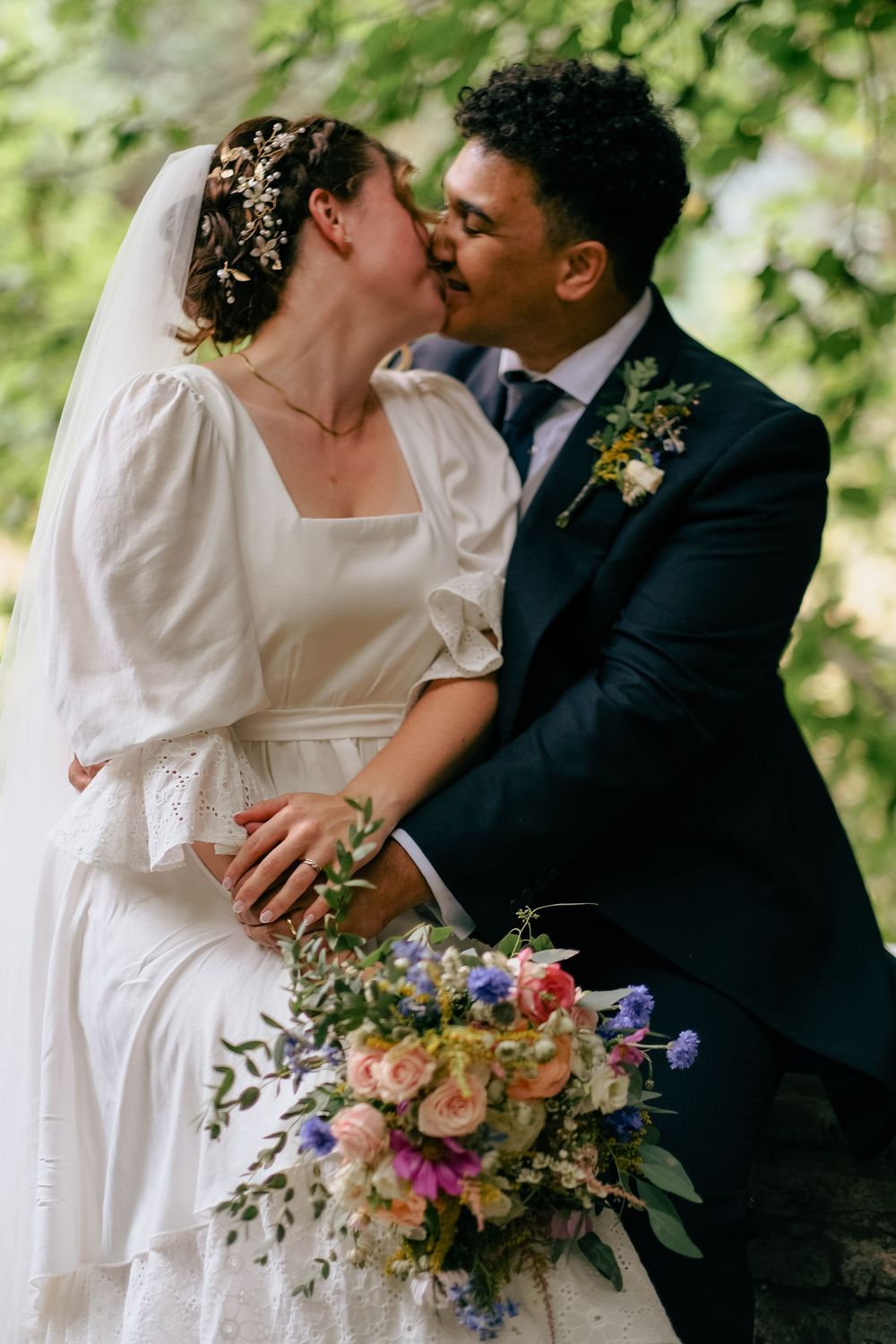 A romantic wedding couple shares a tender kiss outdoors with a beautiful floral bouquet featuring pink and purple blooms.