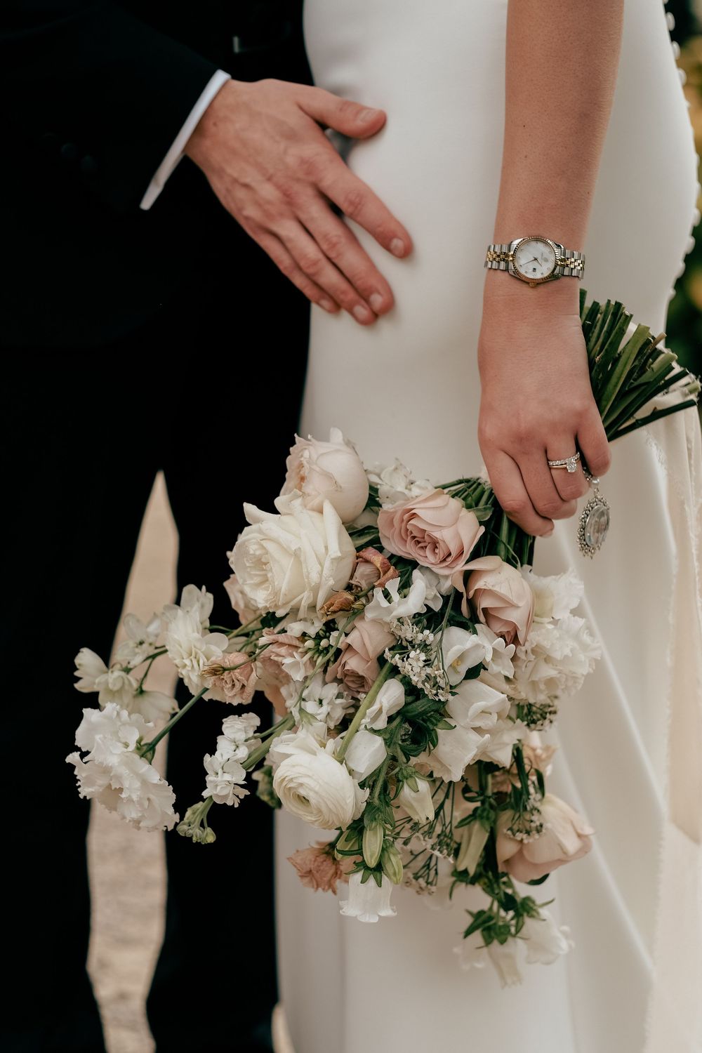 Close-up details of elegant white wedding bouquet and hands with rings against white dress and black suit.