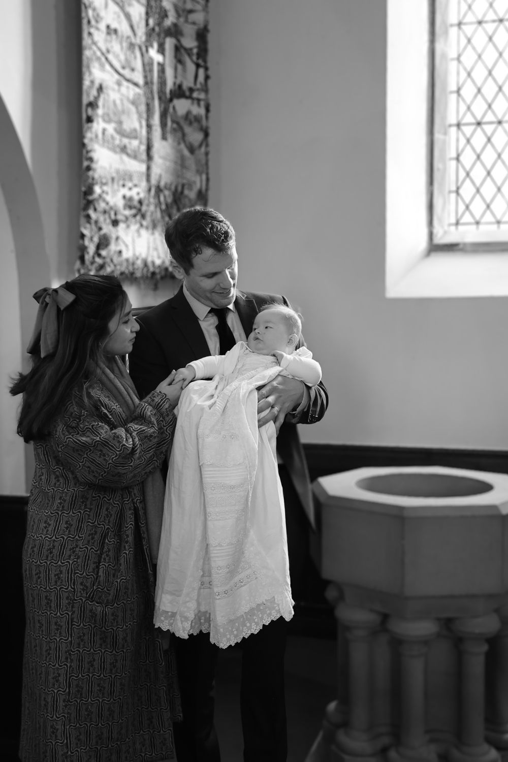 A black and white photograph of a baptism ceremony in a church with a baby in white christening gown.