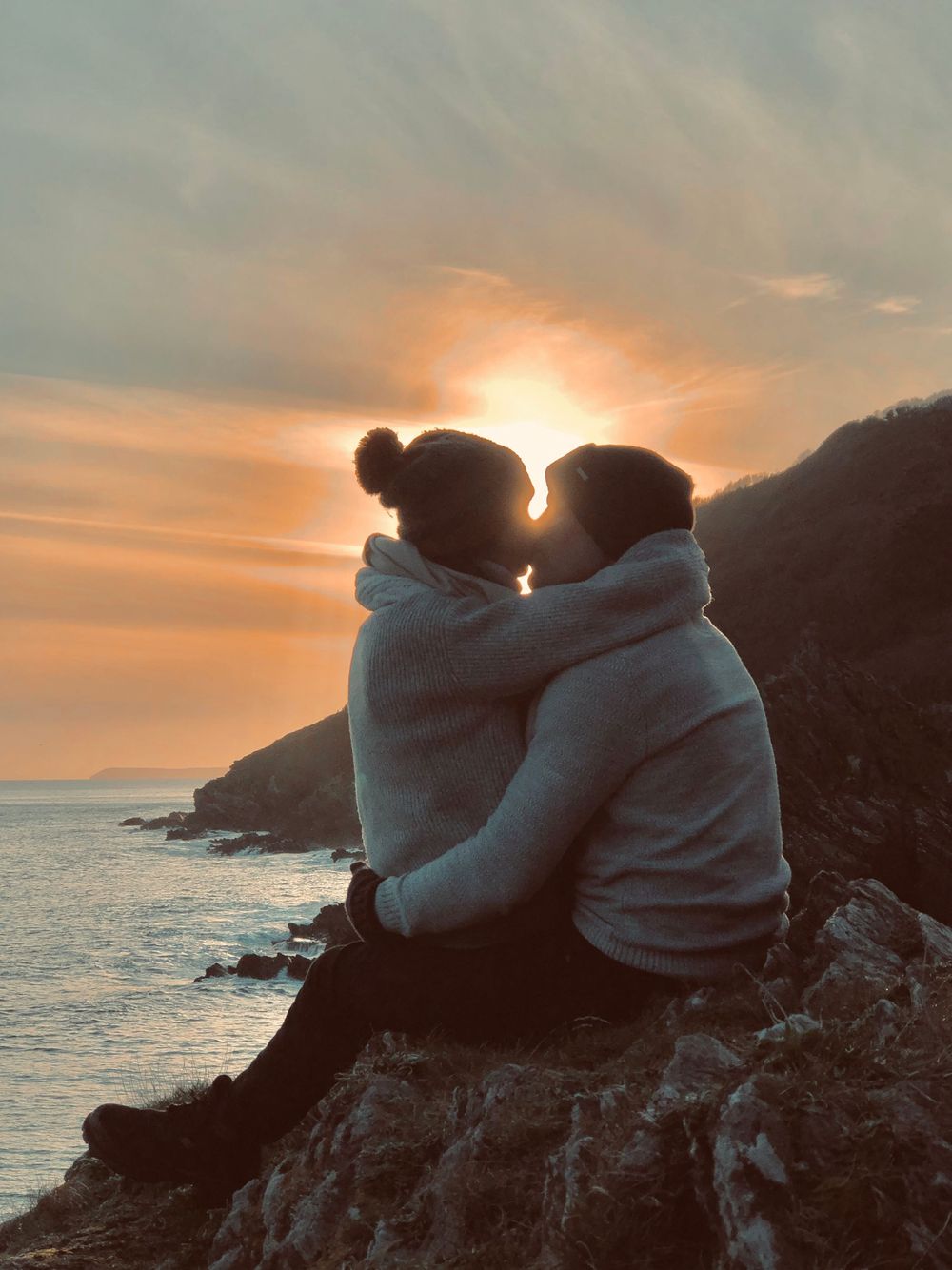 Two people in hooded sweatshirts sitting close together on coastal rocks watching a golden sunset over the ocean.