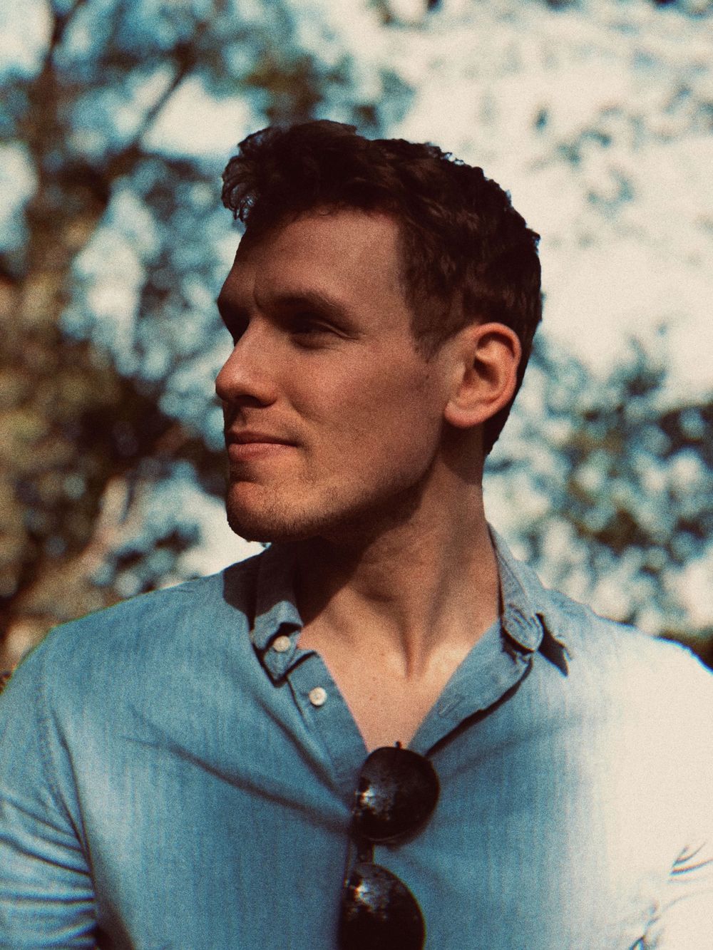 Young man in a light blue button-down shirt with sunglasses hanging from collar poses outdoors on a sunny day.