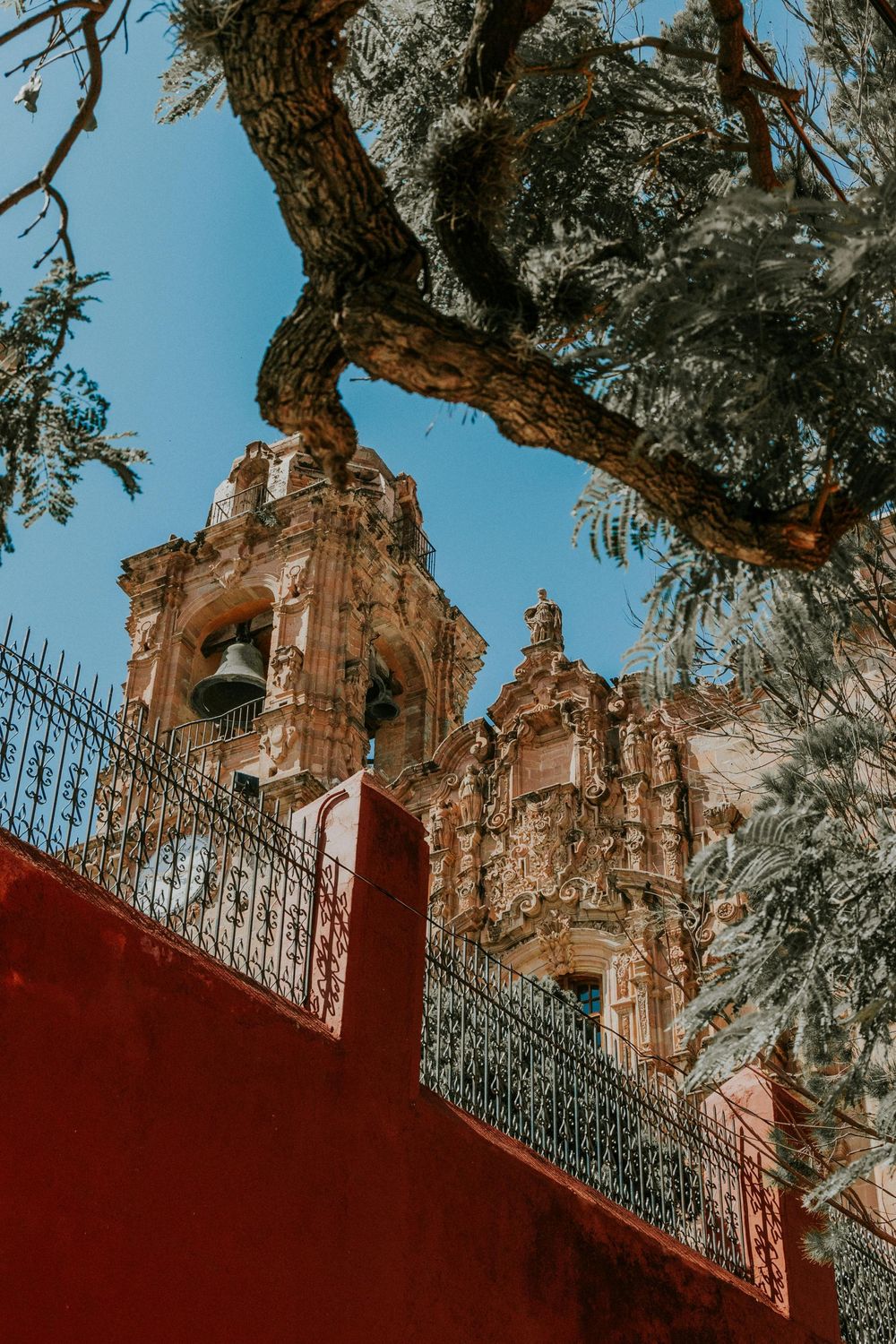 Spanish colonial church bell towers rise above a red wall and ornate iron railing against a blue sky in Mexico.