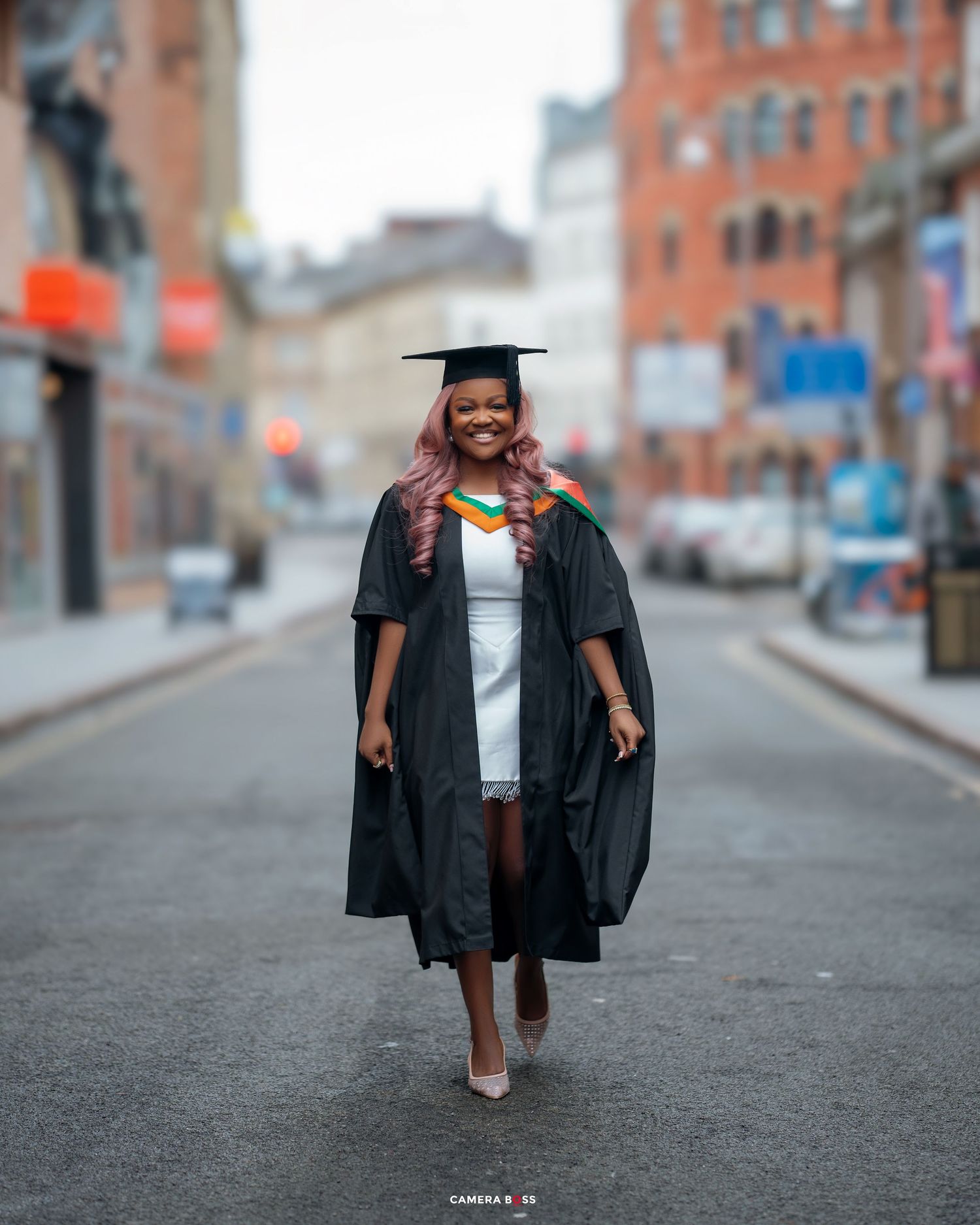 Graduate wearing black cap and gown walking down a city street with red brick buildings in the background.