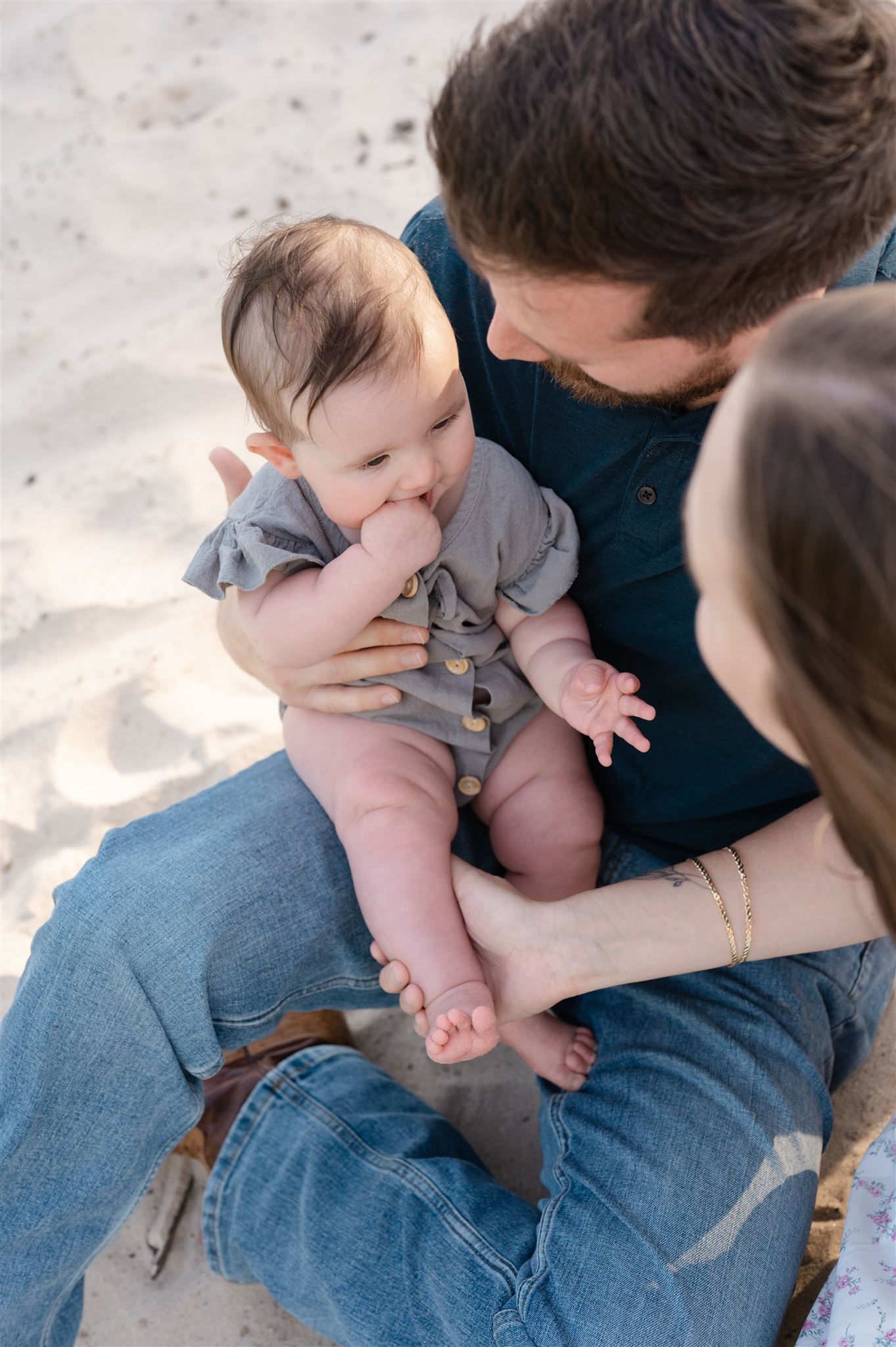A small infant in a gray romper sits on an adult's lap at the beach while sucking their thumb.