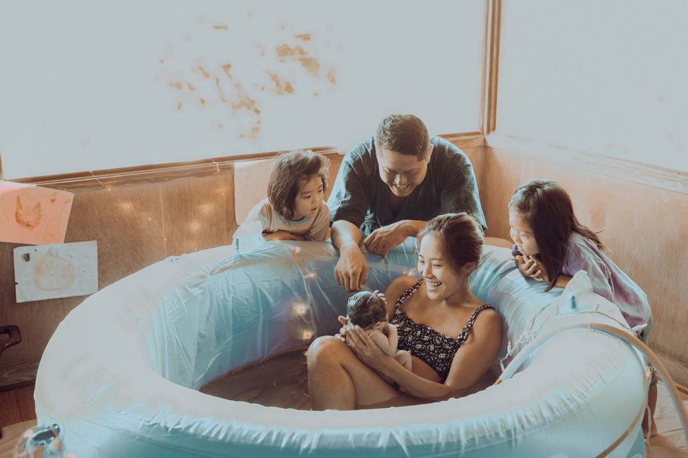 People gathered around a birthing pool supporting and comforting a laboring mother during childbirth. Oahu Newborn Photographer