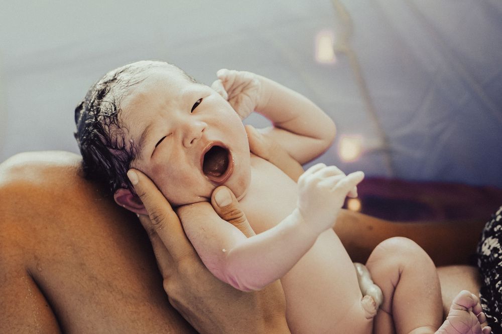 Newborn baby yawning while being gently held during a bath in soft natural lighting. Oahu Newborn Photographer