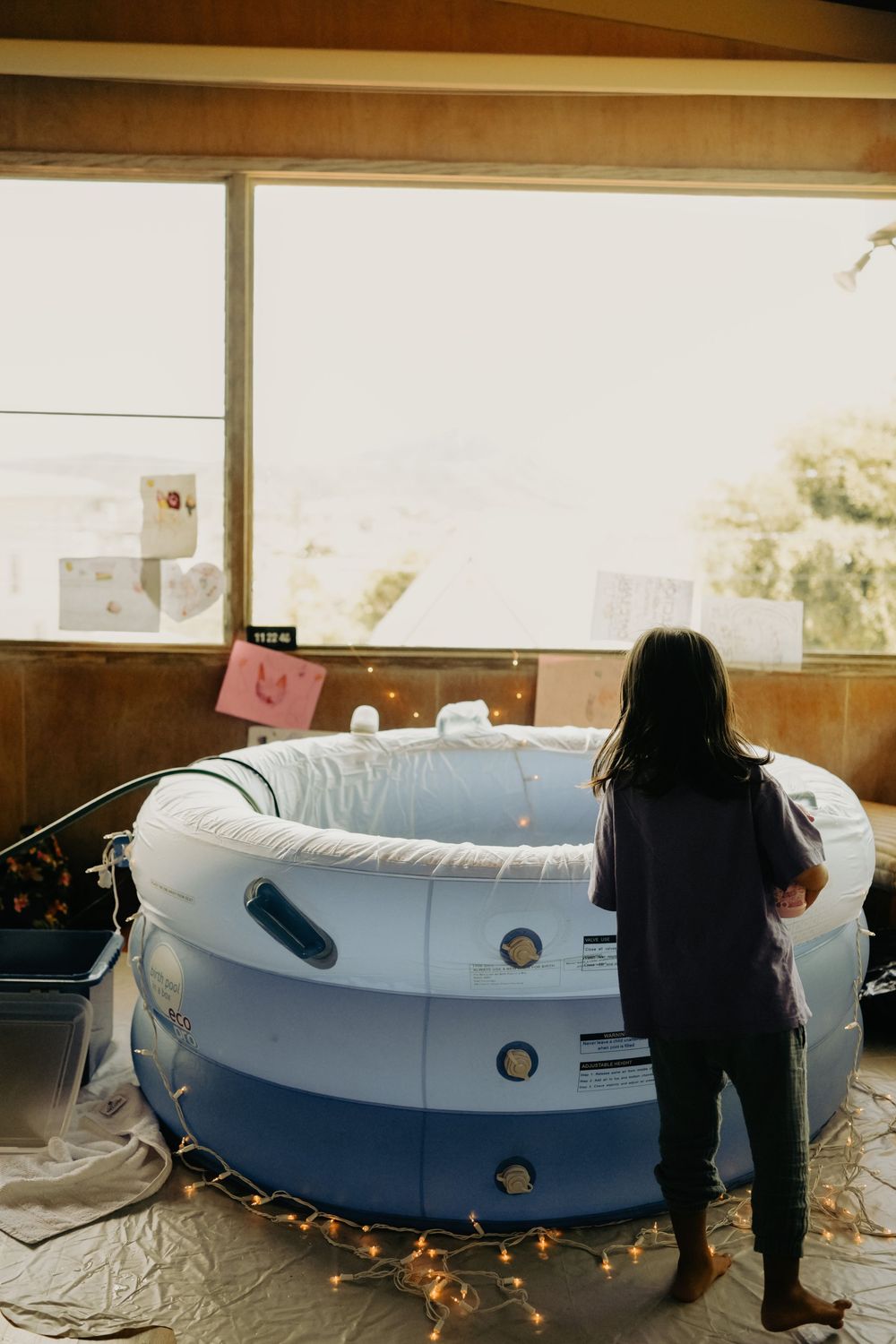 A person stands near an inflated birth pool decorated with string lights in a warmly lit room during golden hour. Oahu Newborn Photographer
