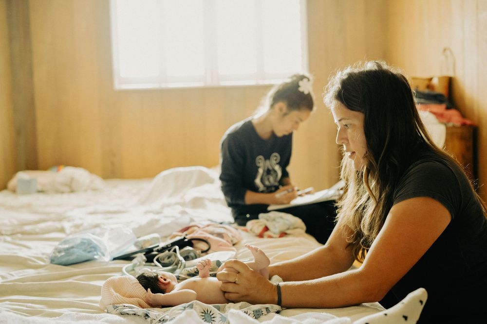Two people sit on a bed in a sunlit room sorting through items spread out on the white bedding. Oahu Newborn Photographer