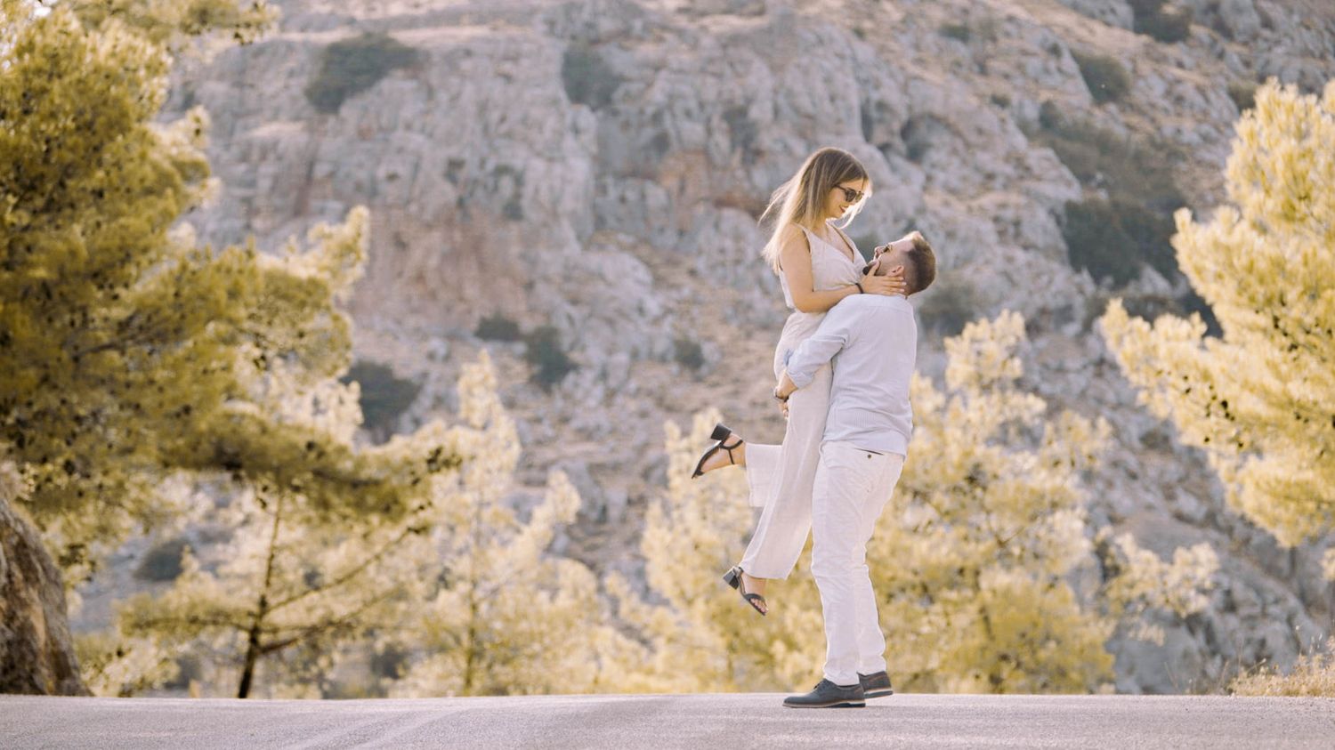 Couple sharing a romantic embrace on a mountain cliff surrounded by pine trees and rocky terrain at sunset.