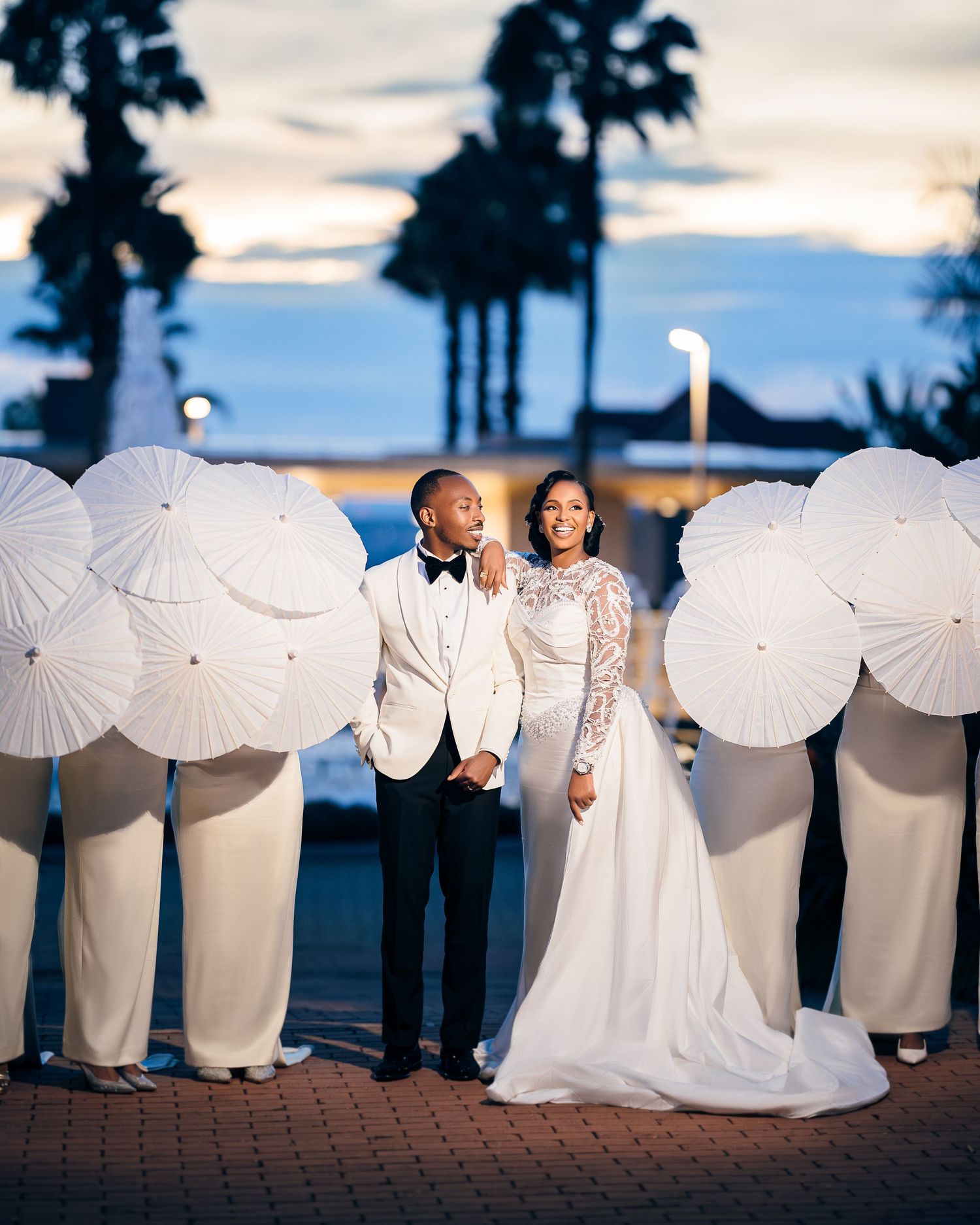 Wedding couple surrounded by guests holding white parasols at sunset with palm trees in the background.