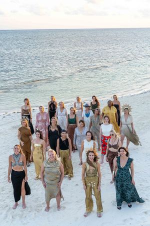 Group photo of women standing together on a white sandy beach at sunset.