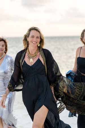 Woman in flowing black dress walks along beach wearing statement necklace.