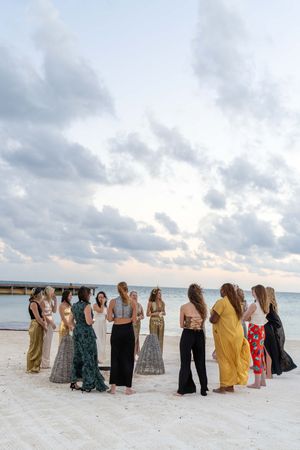 Group of friends in flowing dresses gathered on a pristine beach at sunset with cloudy skies in the background.