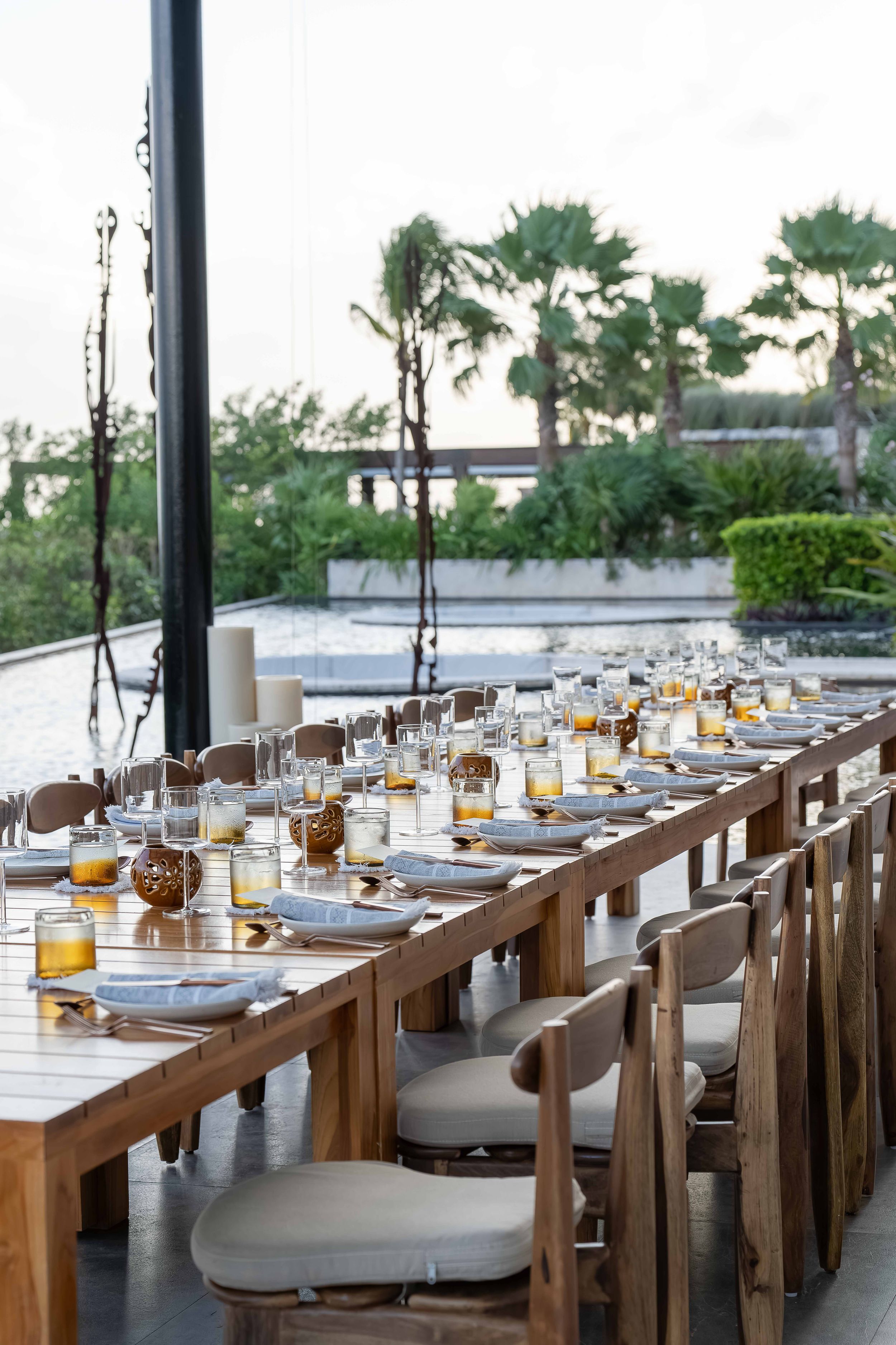 Elegant long dining table set with glowing candles and place settings overlooking palm trees at dusk.