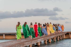 Group walking along wooden pier wearing colorful flowing dresses against ocean backdrop.