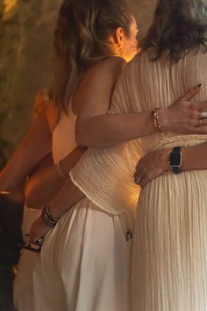 Two people in white dresses embrace during a warmly lit celebration event.