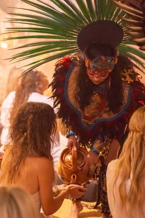A performer in an elaborate feathered headdress and mask interacts with guests at a tropical celebration.