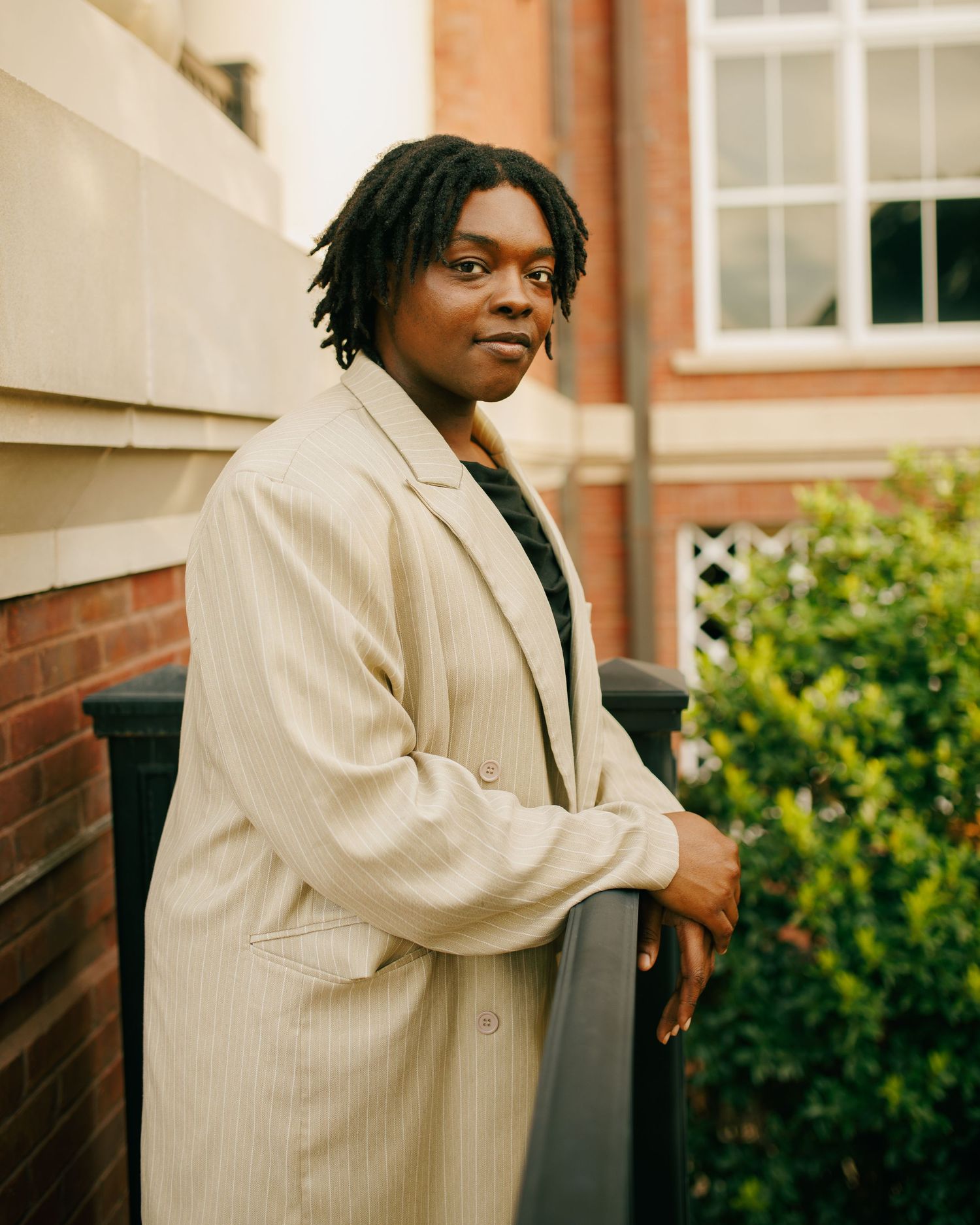 Person in beige pinstripe blazer poses casually against brick building exterior.