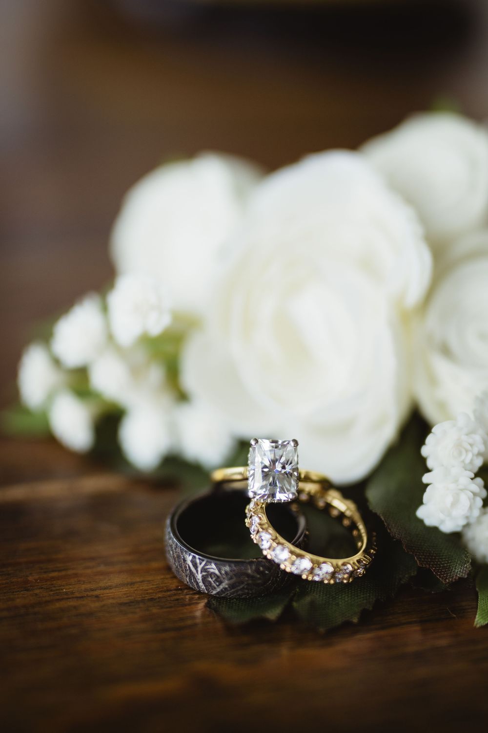 Wedding rings and white roses displayed on dark wooden surface with elegant engagement ring and diamond eternity band.