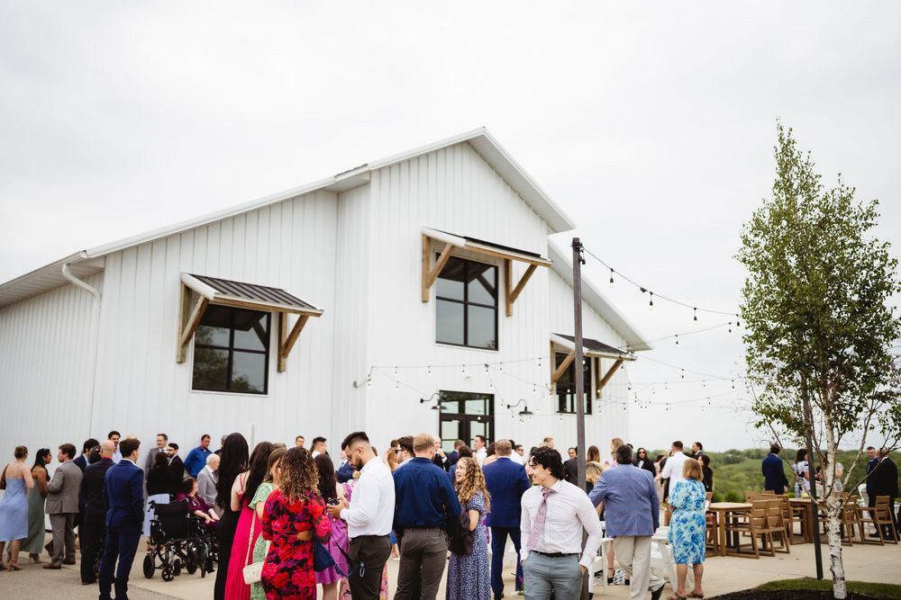 A crowd gathers outside a modern white barn venue with string lights and wooden window awnings.