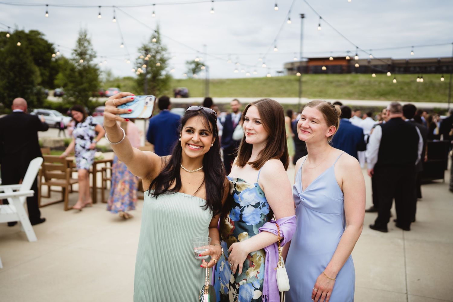 Three guests in pastel dresses celebrate together at an outdoor reception venue with festive string lights overhead.