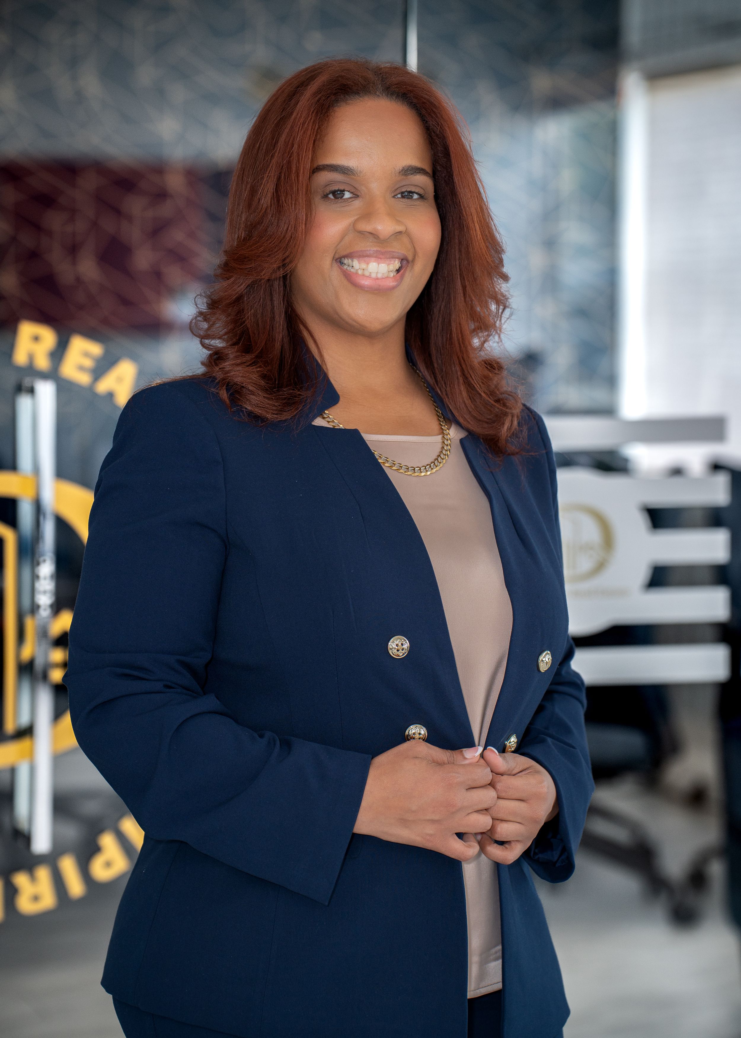 Professional portrait of a person wearing a navy blue blazer and beige top in an office setting with signage in background.