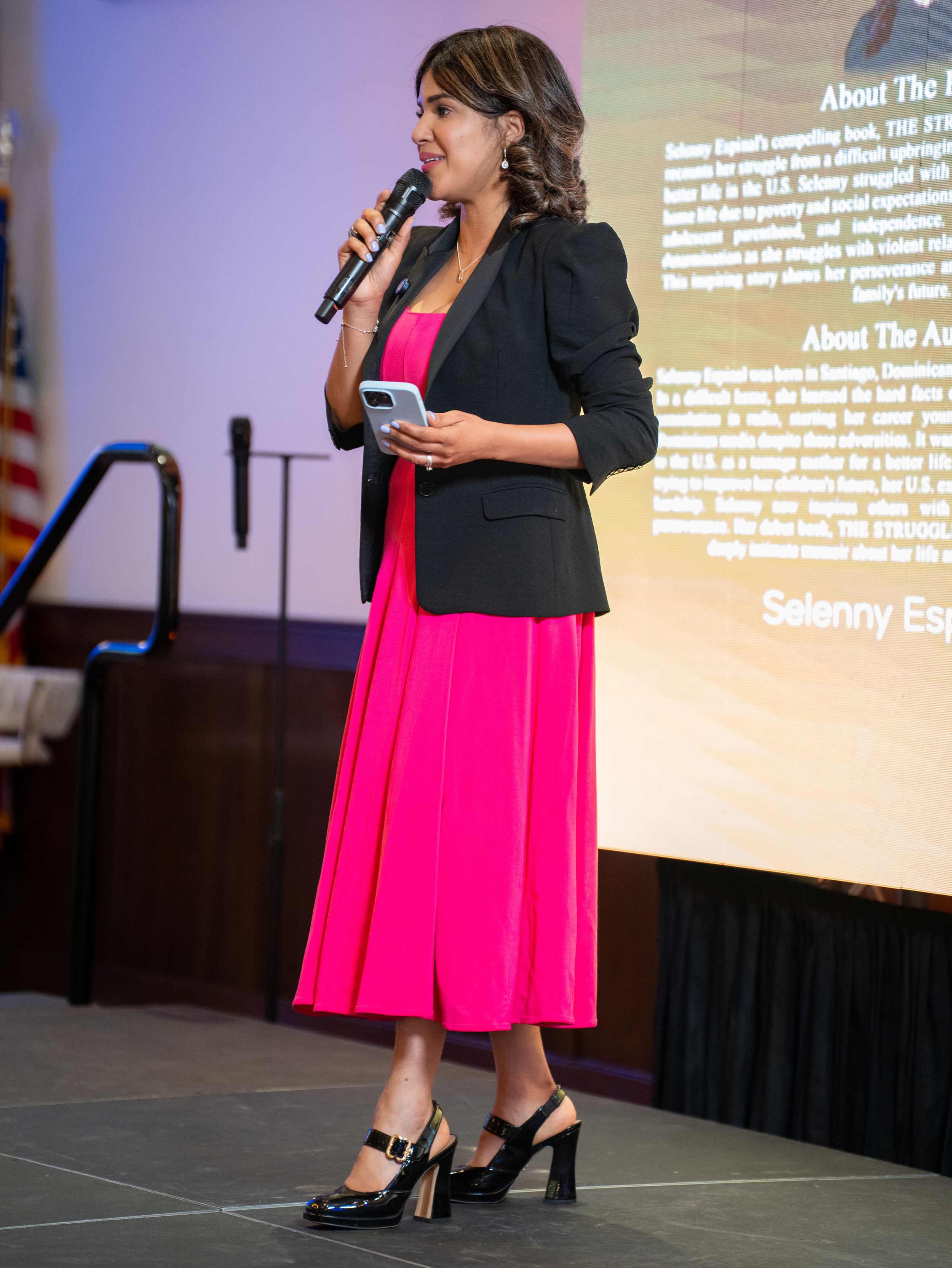 Presenter wearing a black blazer and bright pink skirt speaks into microphone on stage with presentation screen behind.