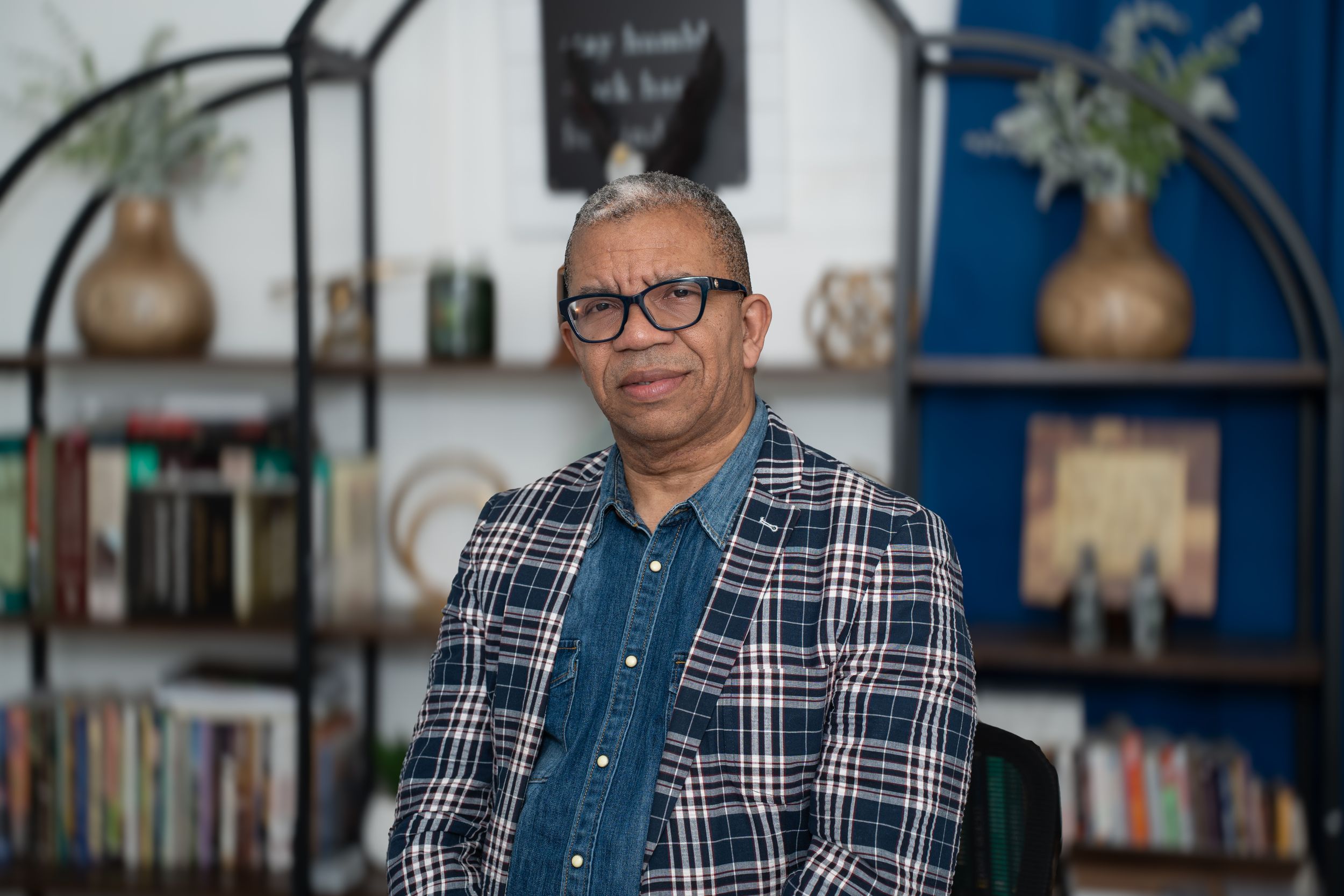 Person wearing glasses and a plaid blazer sits in front of blue bookshelf with decorative vases.
