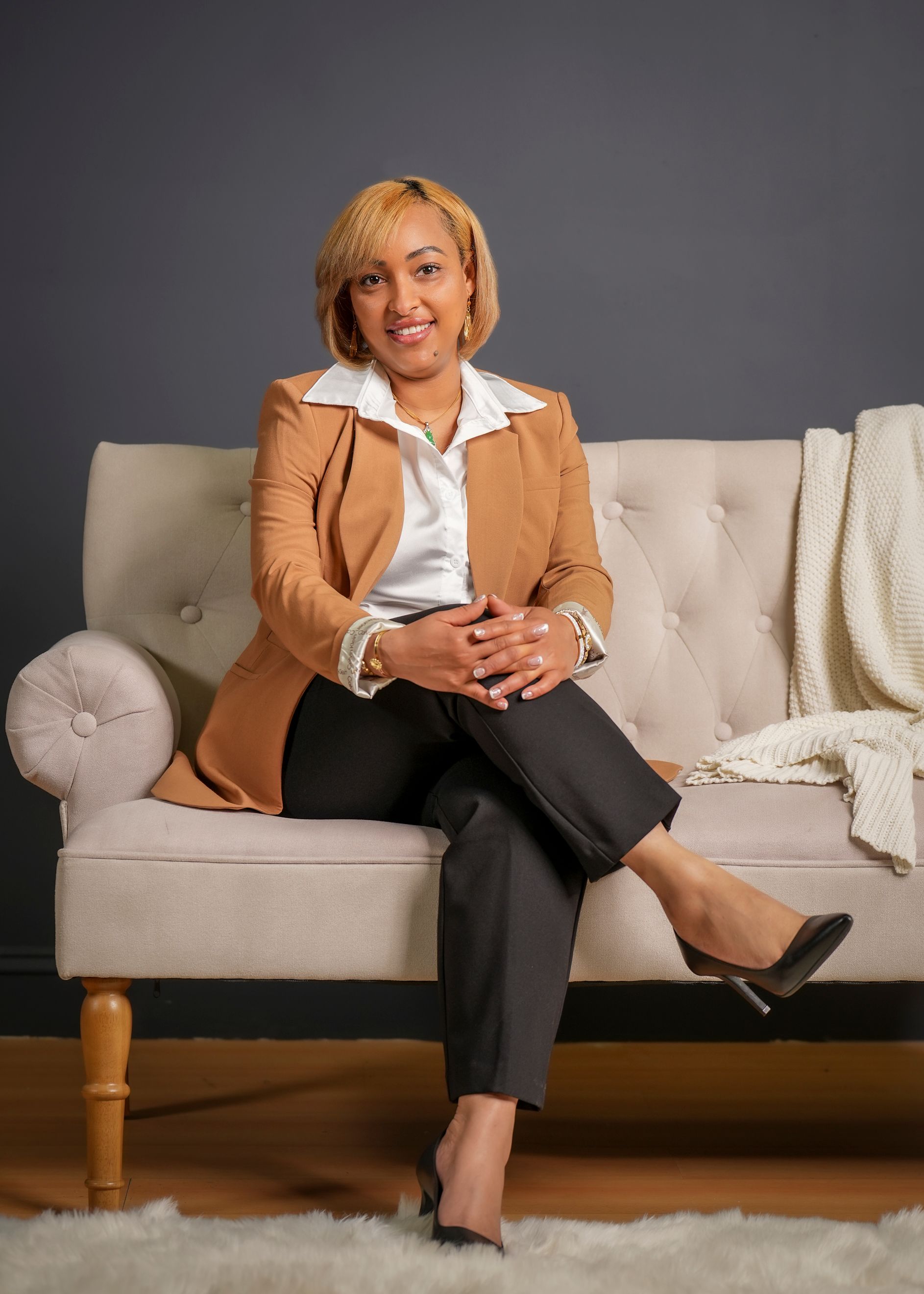 Professional wearing a tan blazer and white shirt sits on a cream-colored sofa against a dark gray wall.