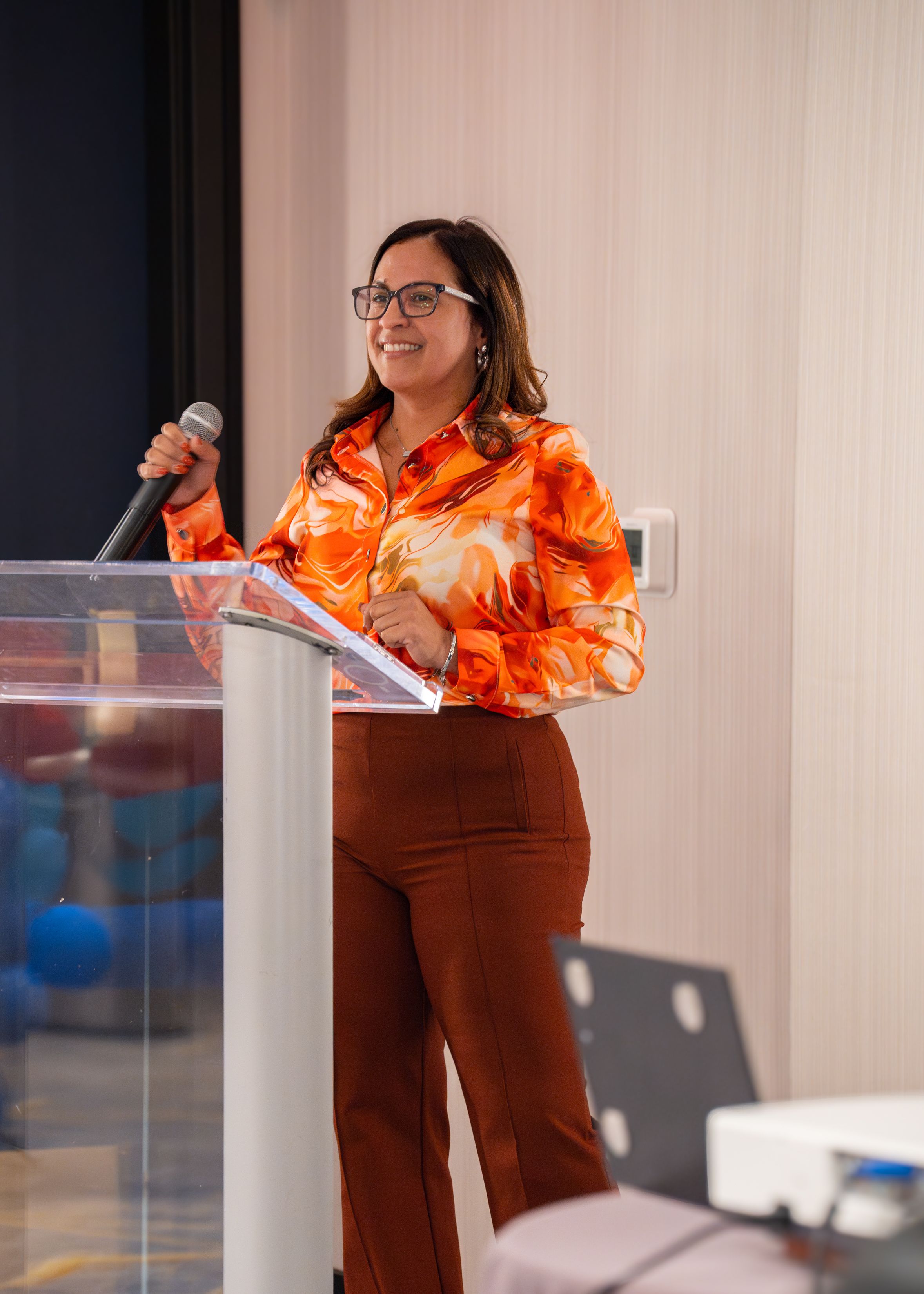 Someone wearing an orange patterned blouse and brown pants speaks at a podium with a microphone during a presentation.