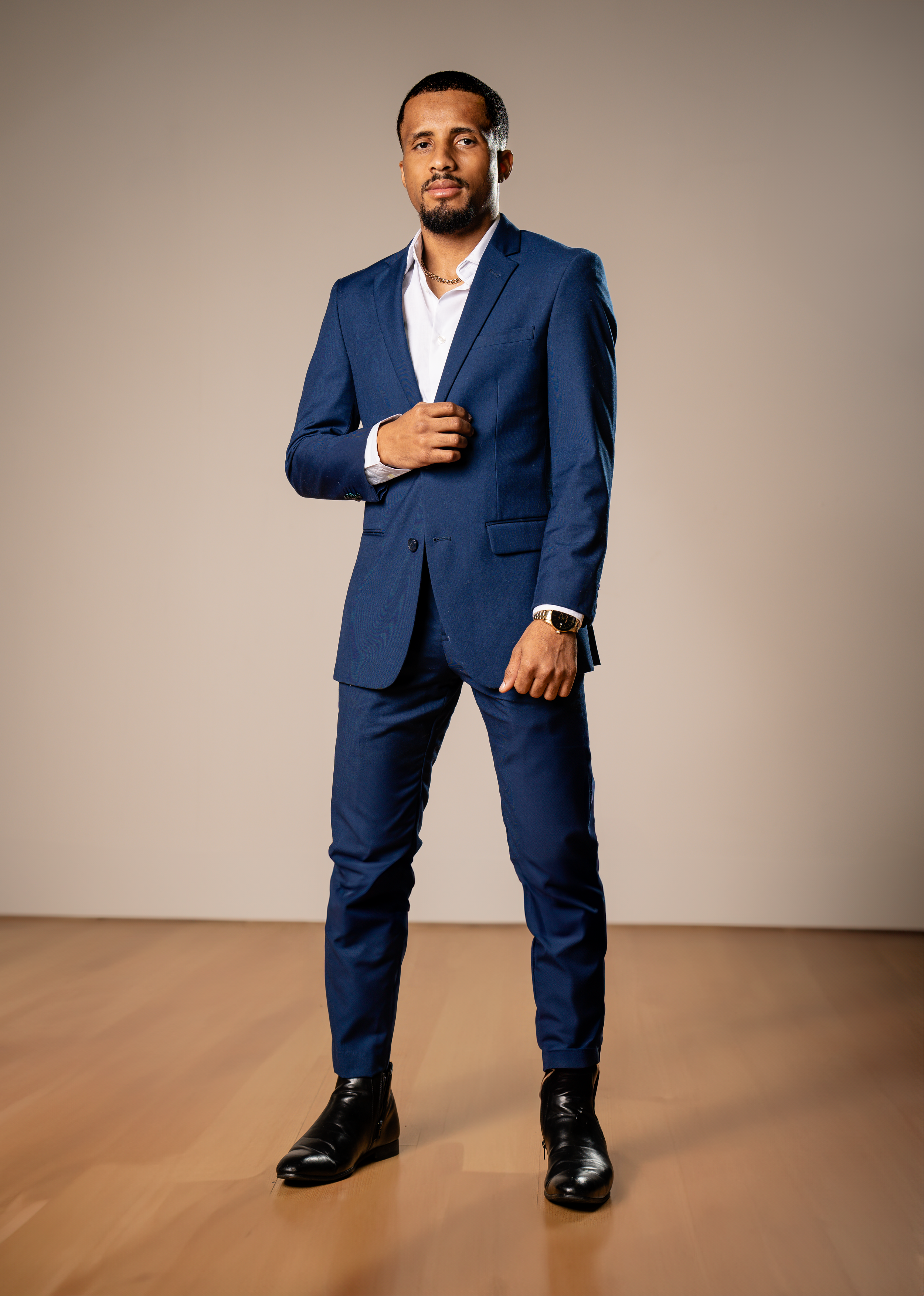 A person poses in a tailored navy blue suit with white dress shirt and black shoes against a neutral background.
