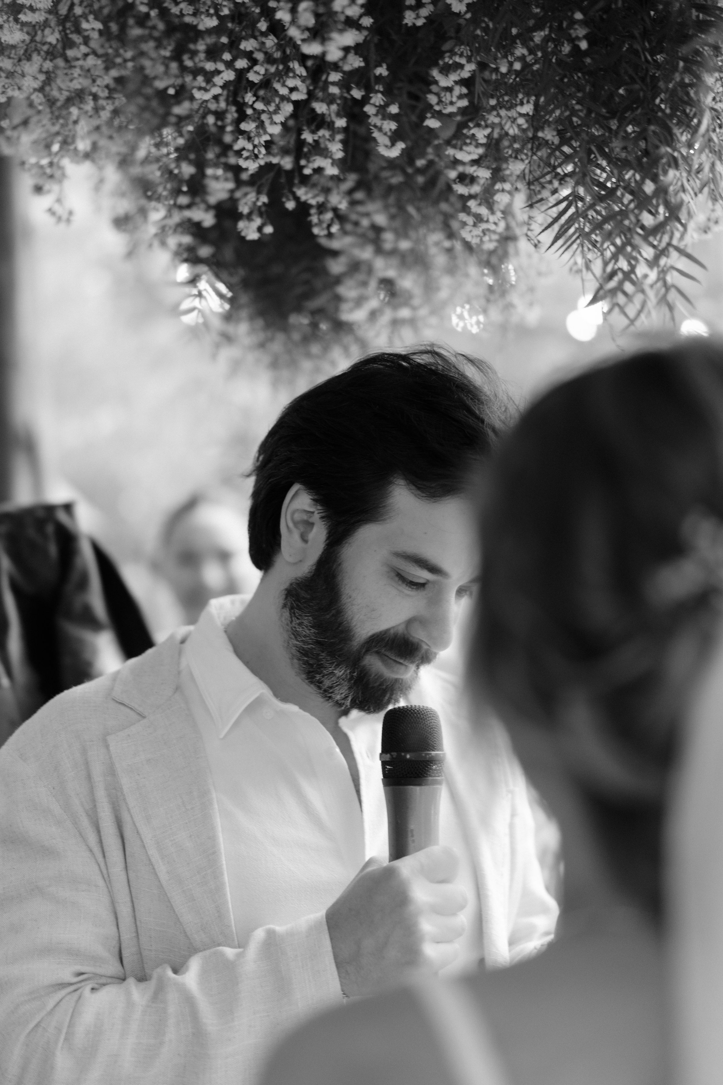 A black and white portrait of someone speaking into a microphone under hanging floral decorations.