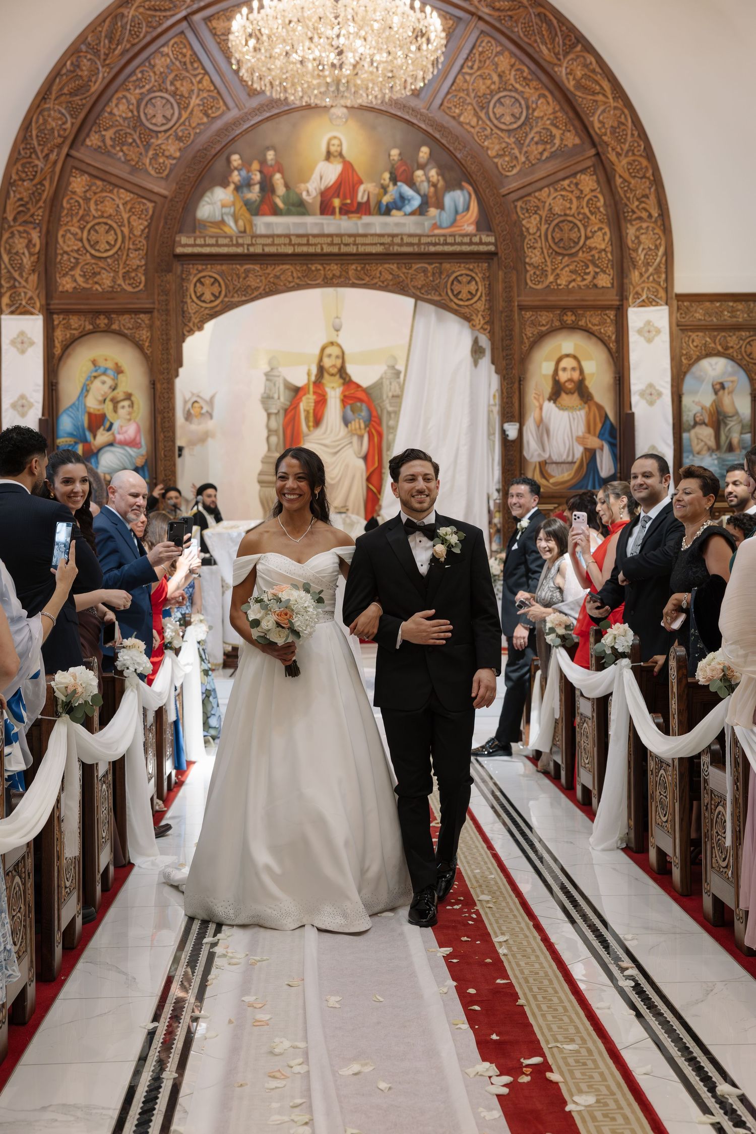 A romantic outdoor wedding ceremony takes place under grand oak trees with decorative lanterns and red flowers lining the aisle.