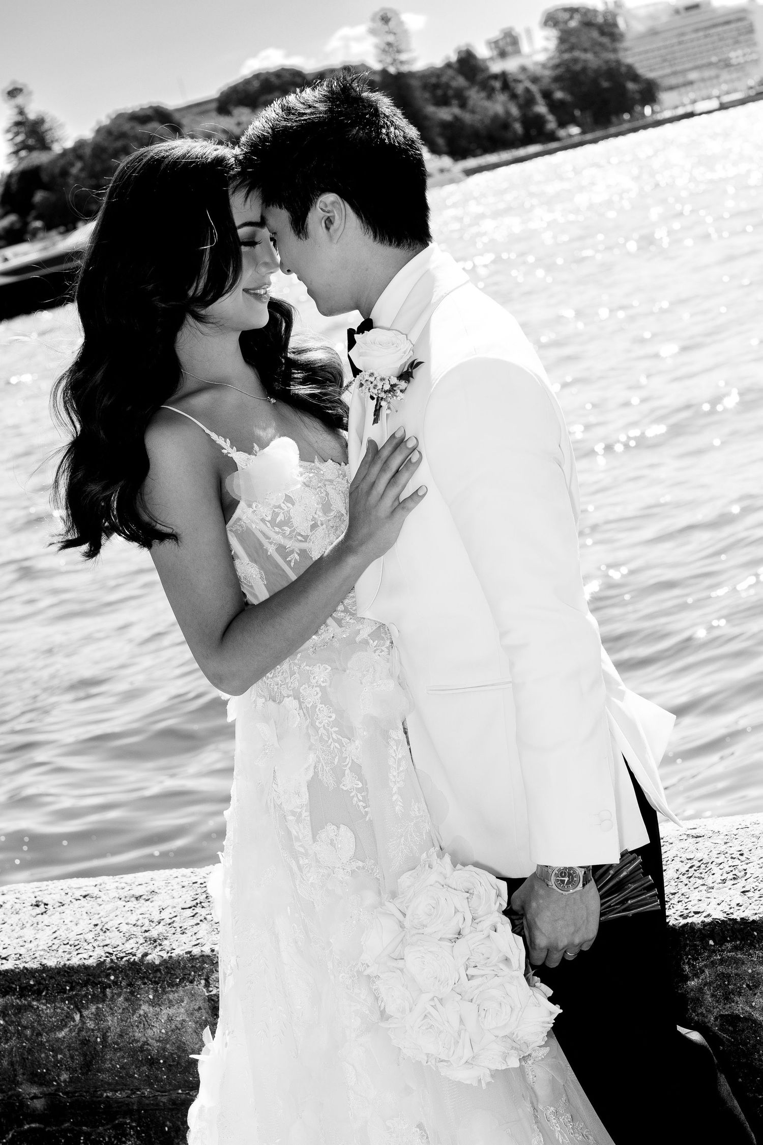 Black and white wedding portrait of a couple in elegant attire embracing by a waterfront on their special day.