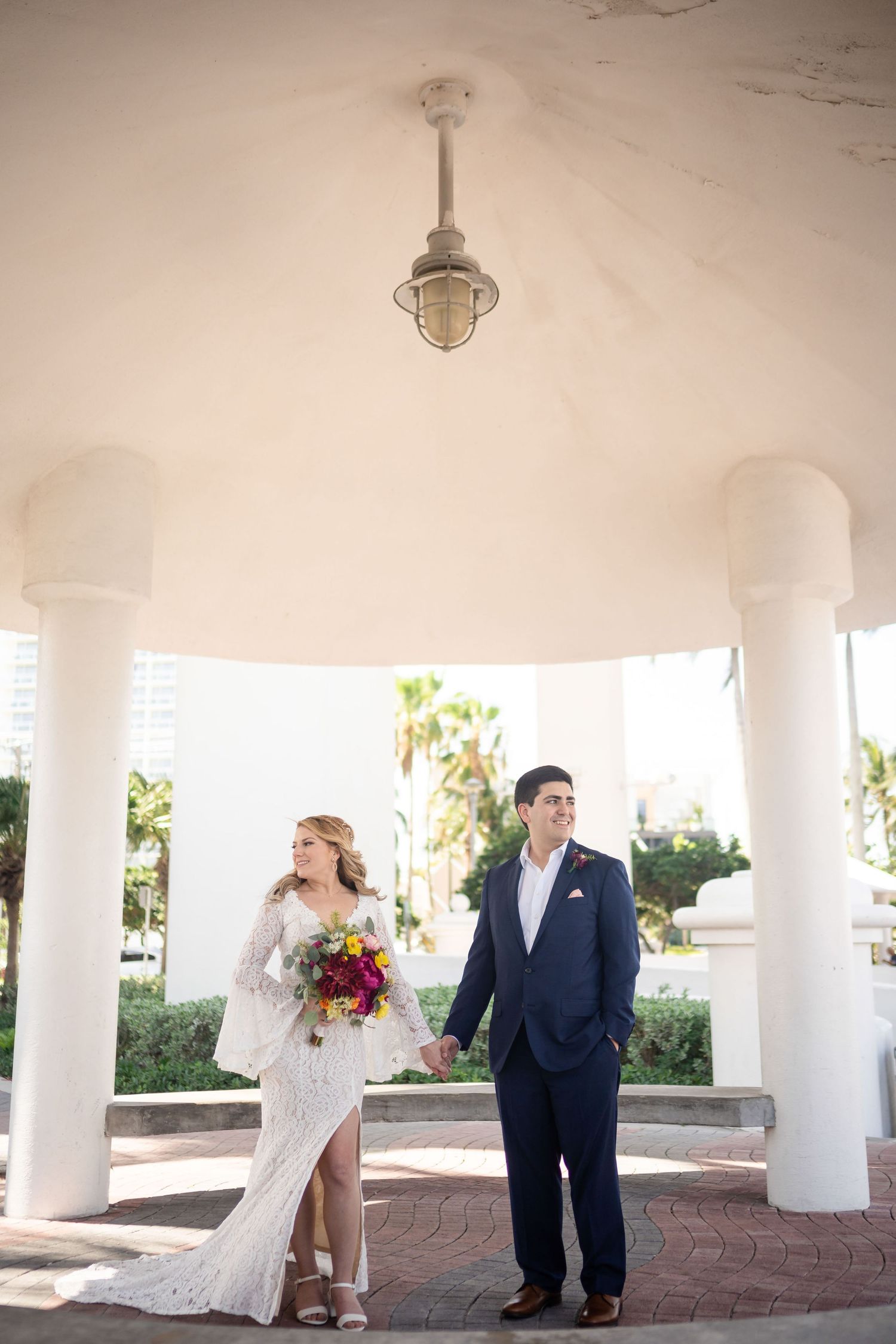 A couple poses under a white columned gazebo during their wedding ceremony with palm trees visible in the background.