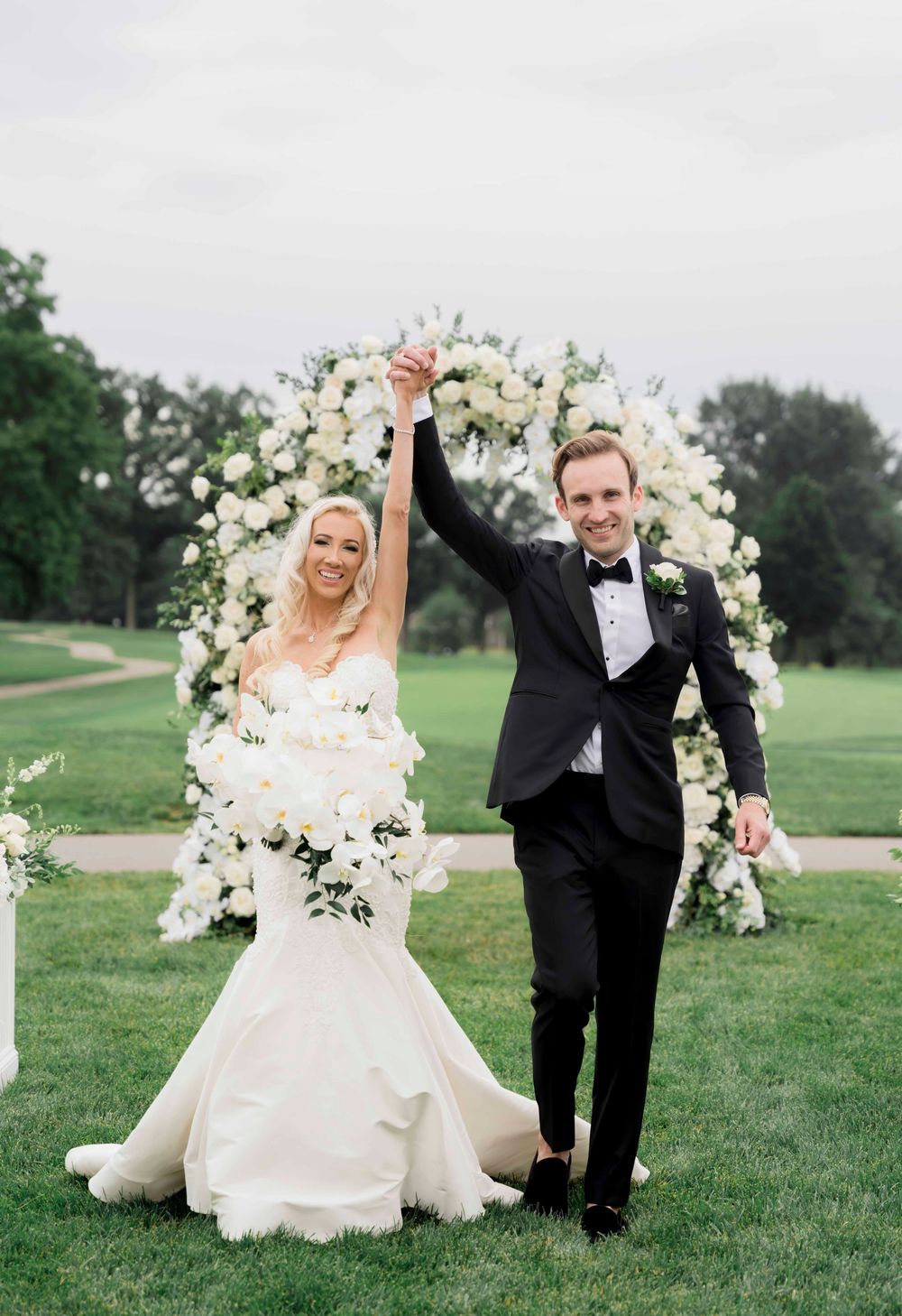 A newly married couple celebrates under a lush white floral arch on a golf course during their wedding ceremony.