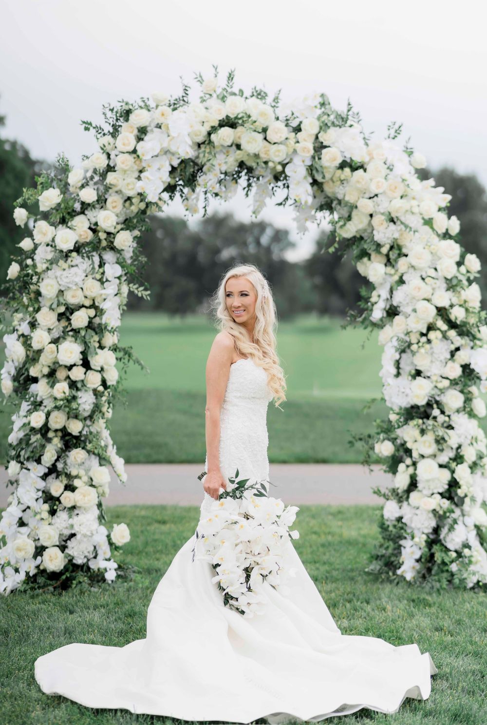 Beautiful white floral wedding arch with roses and greenery frames an elegant bridal portrait on a lush grass lawn.