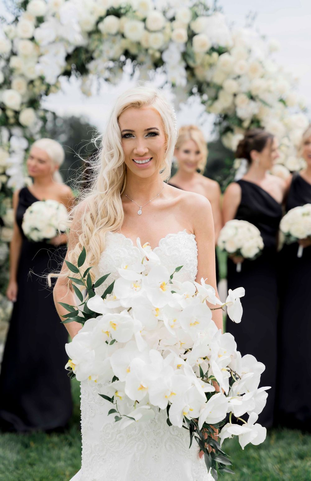 A stunning white wedding bouquet featuring cascading orchids against a floral arch backdrop at an outdoor ceremony.