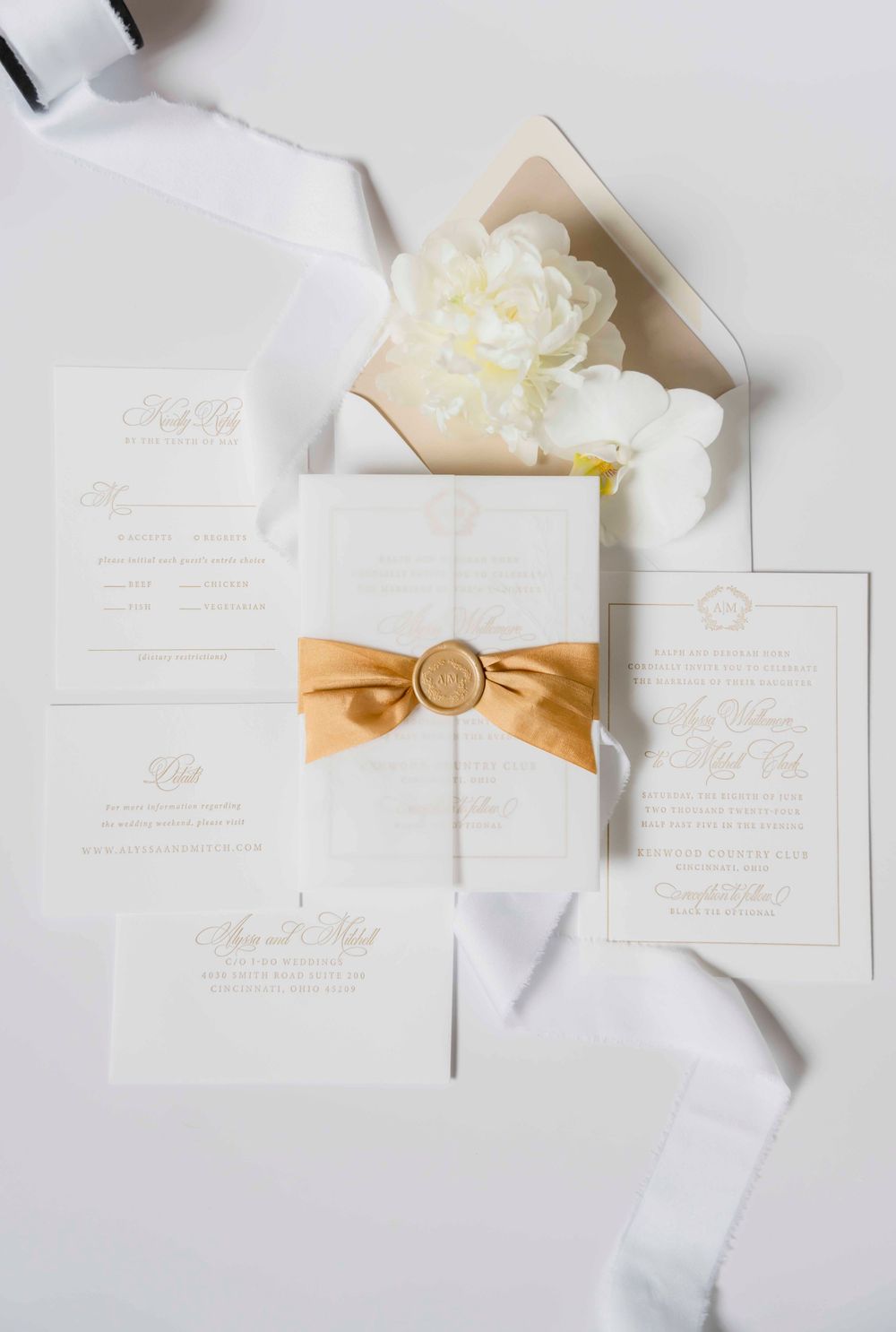 Elegant white wedding invitation suite with gold wax seal and ribbon on a light background with floral accents.