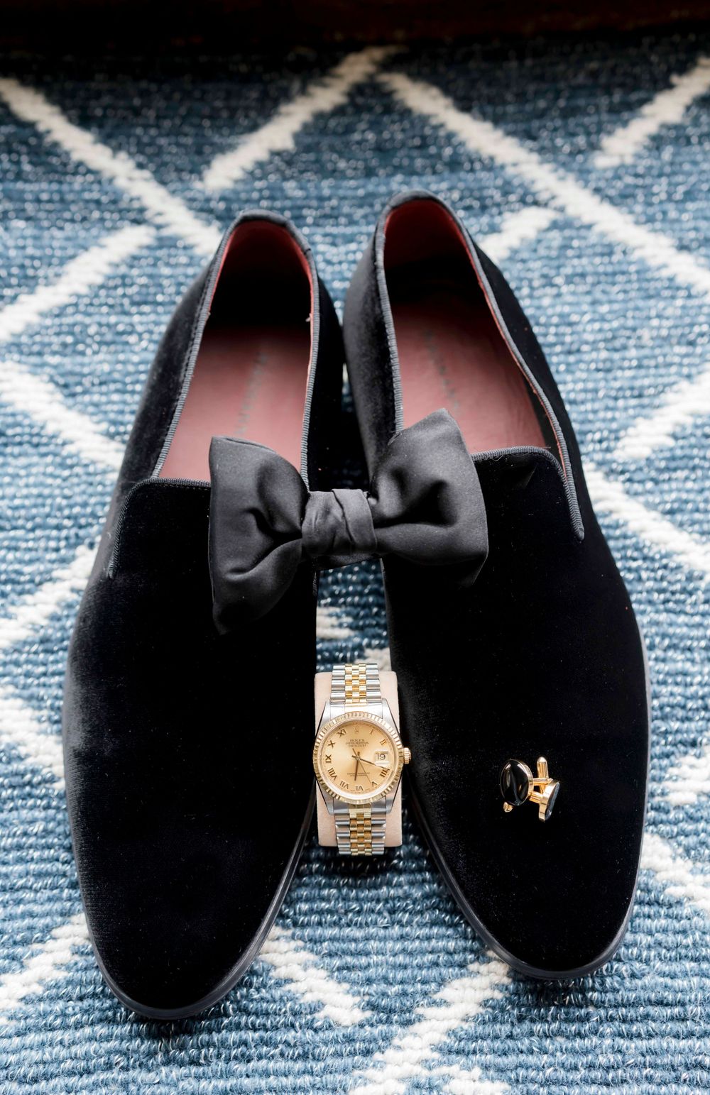 Black velvet loafers with bow ties displayed with a gold watch on a geometric patterned rug.