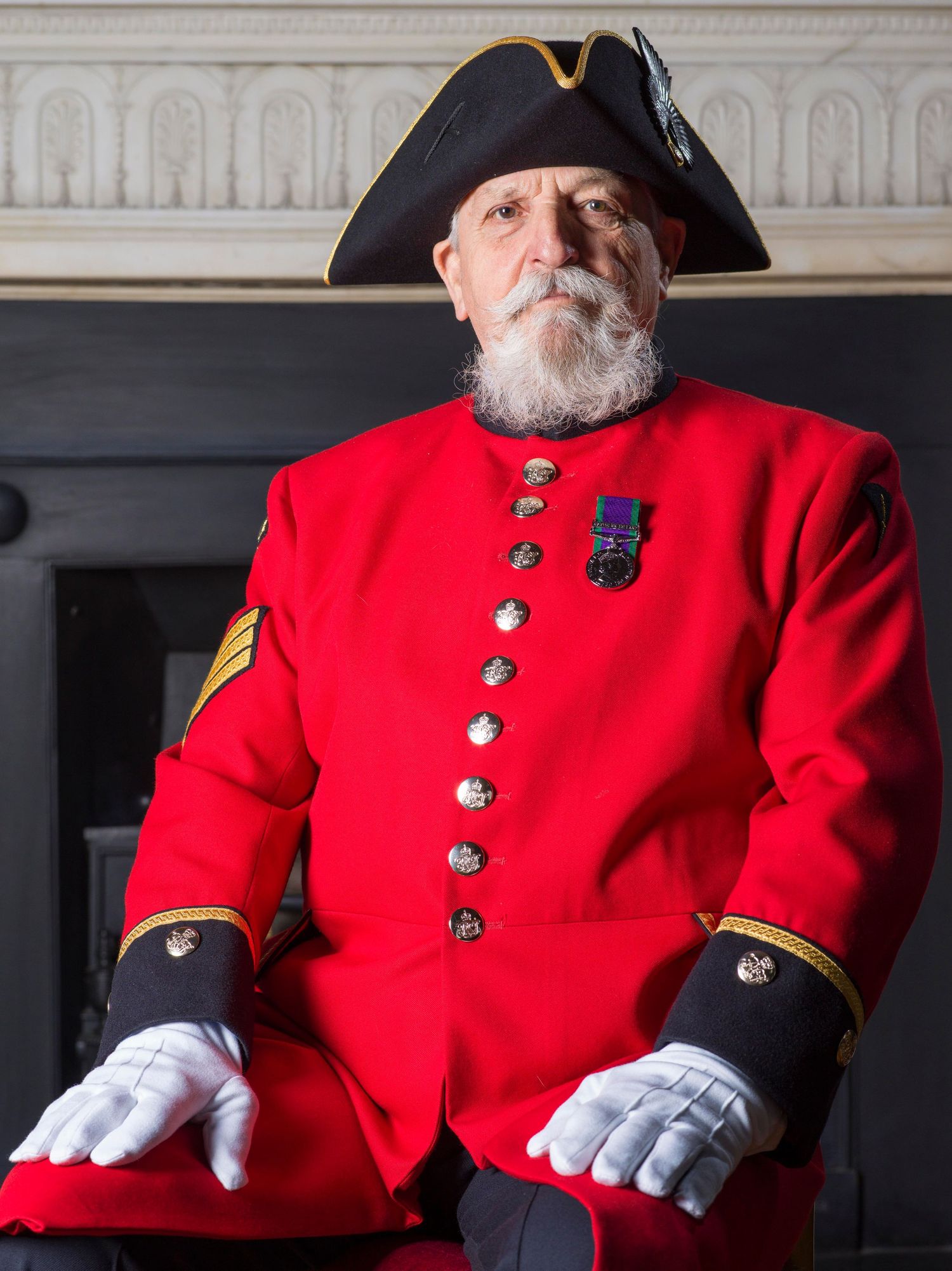 A Chelsea Pensioner in traditional red military uniform with tricorn hat and medals sits formally against an ornate background.