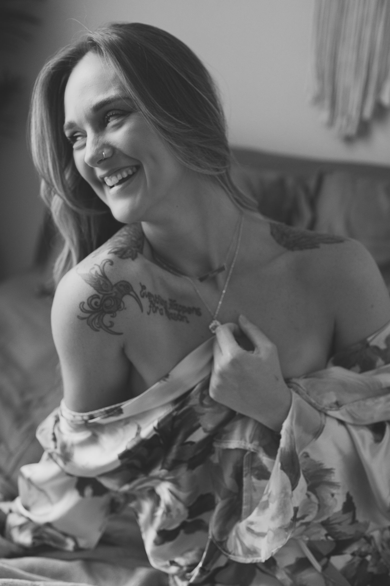 Black and white portrait features a natural candid smile and visible shoulder tattoo while wearing off-shoulder clothing.